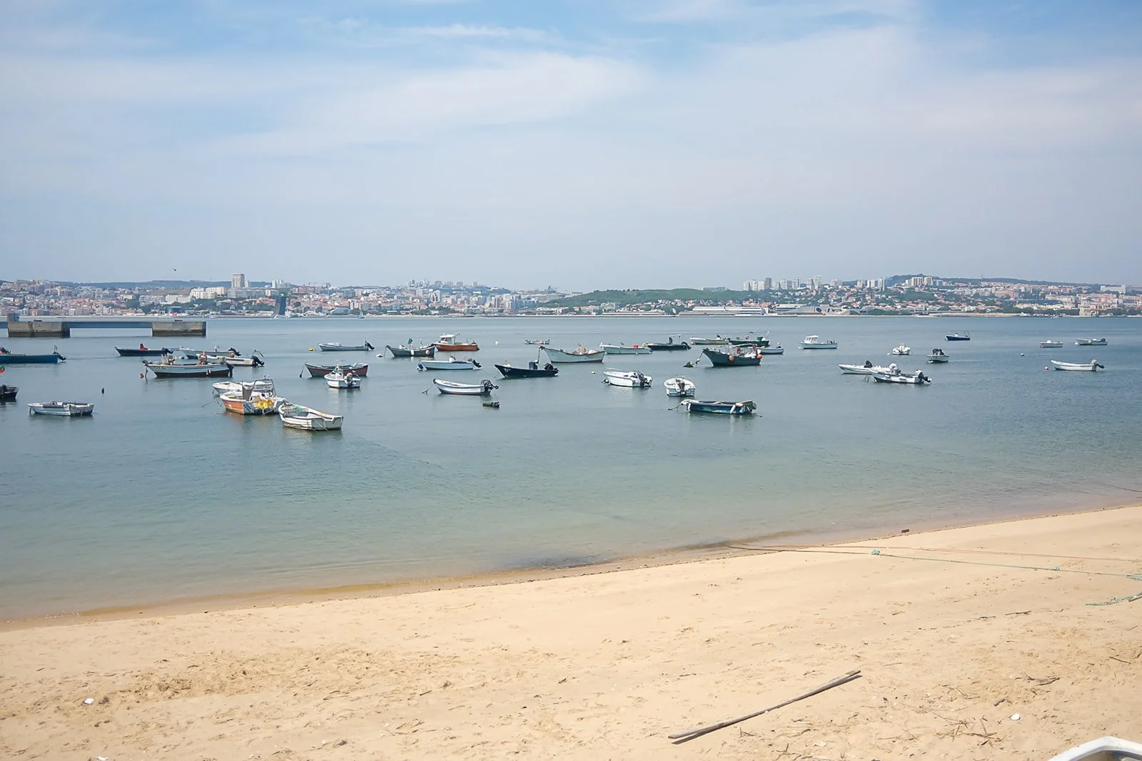 A scenic view of a harbor with numerous boats anchored in calm water, a sandy beach in the foreground, and a city skyline with various buildings in the distance under a partly cloudy sky.