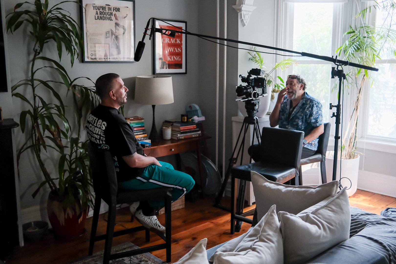 A man being interviewed on a video set in a living room with large windows, potted plants, a bookshelf, a floor lamp, and framed posters on the wall.
