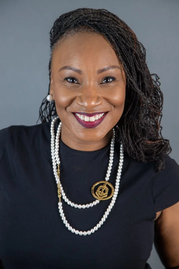A woman with shoulder-length dreadlocks, smiling, wearing a black top, pearl necklace, and gold-colored jewelry, with a gray background.