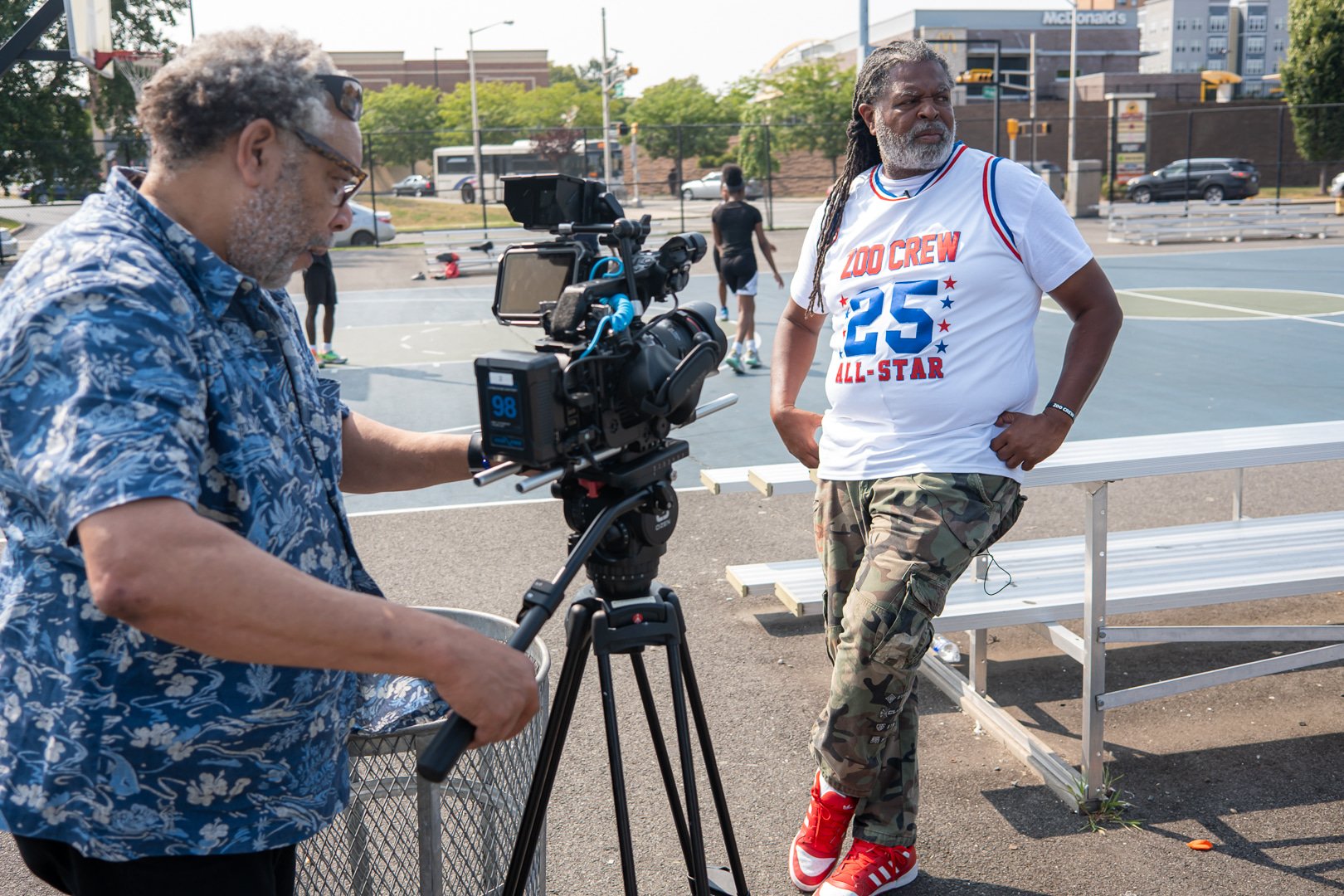 A man with gray hair and glasses is filming with a camera on a tripod while another man with dreadlocks and a beard stands nearby on a basketball court, wearing a white t-shirt that says "Zoo Crew 25 All-Star" and camouflage pants.
