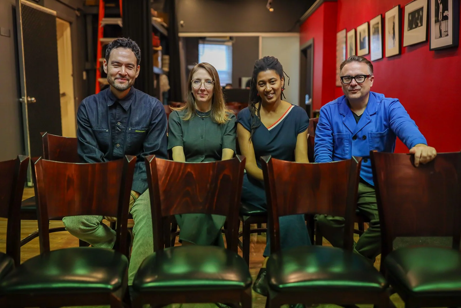 Four people sitting in a row of chairs in a cozy restaurant or cafe with a red accent wall and framed pictures. They are dressed casually and smiling at the camera.