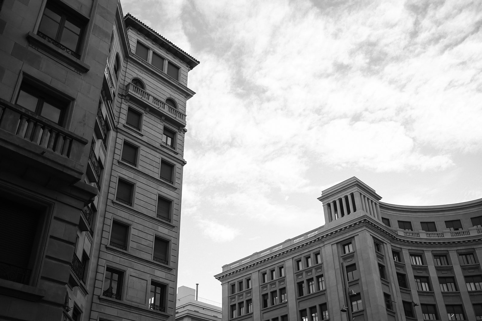 Black and white photo of tall, ornate buildings with classical architectural details, viewed from the street with a cloudy sky overhead.
