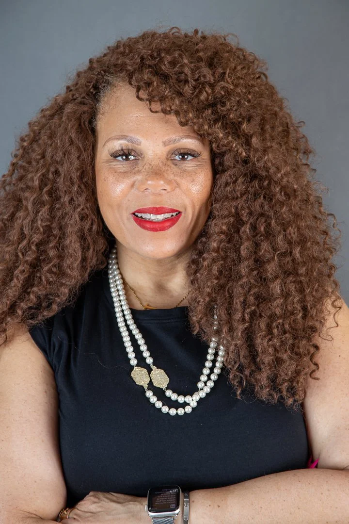 A woman with curly red hair wearing a black top, red lipstick, a pearl necklace, and a smartwatch on her wrist, smiling at the camera against a gray background.