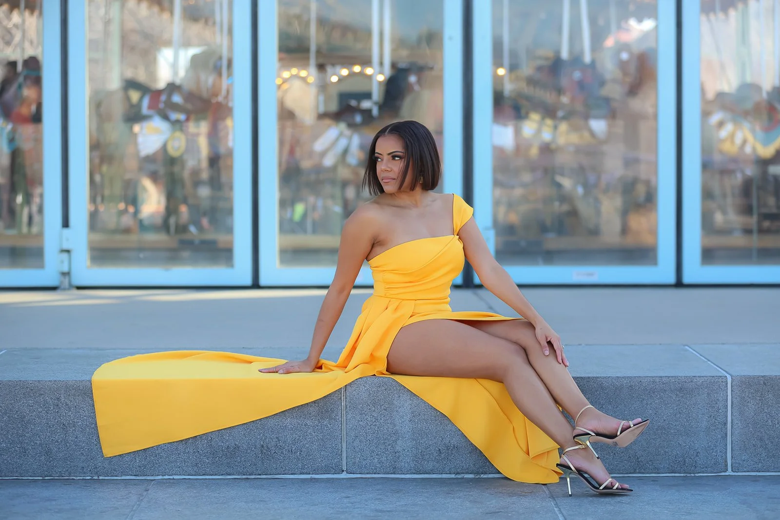 Woman in a yellow dress sitting on a ledge with a glass building in the background.