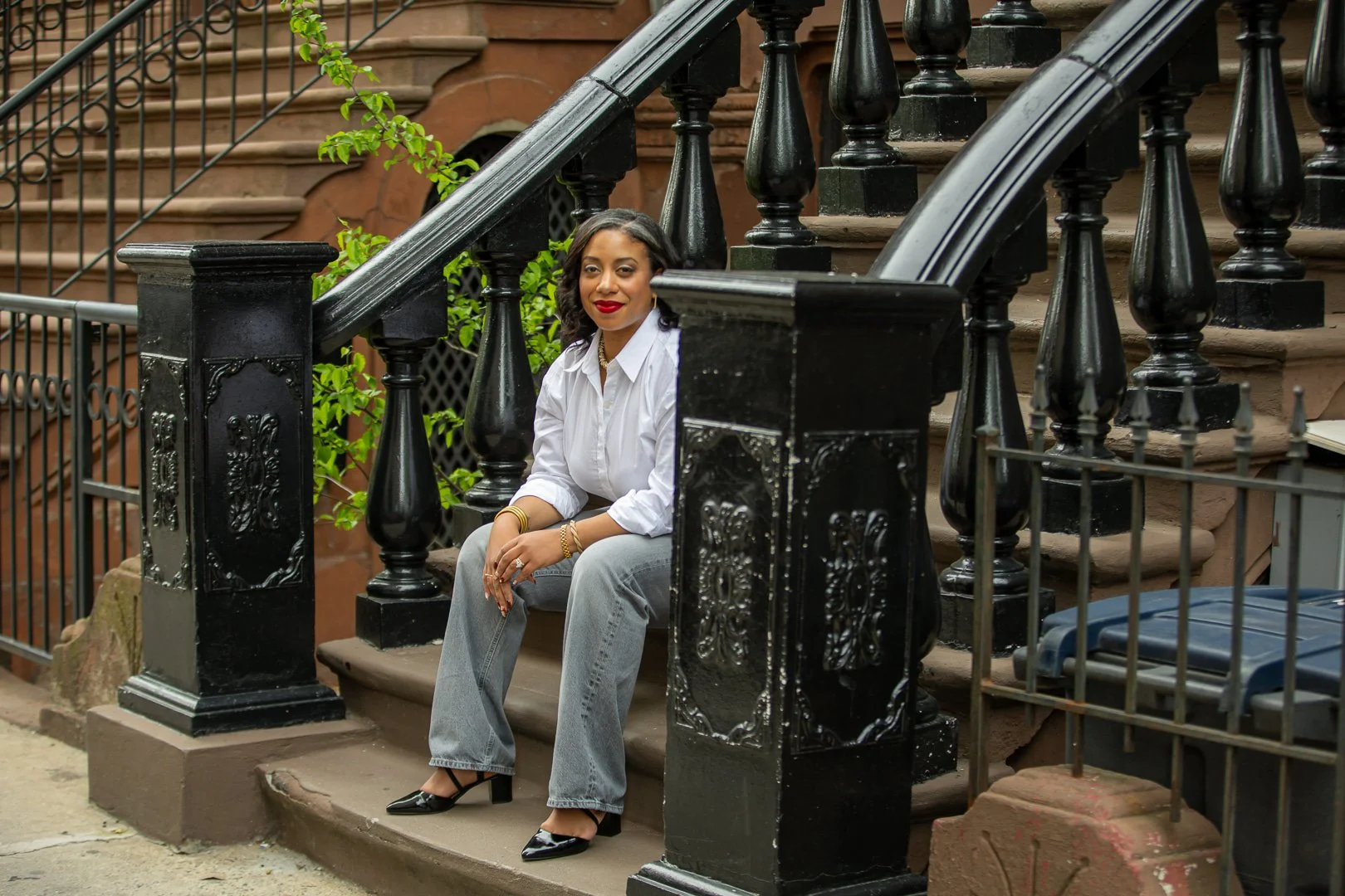 A woman sitting on brown stairs outside a building with ornate black iron railings and green plants around. She is wearing a white shirt, gray pants, black heels, red lipstick, and gold jewelry, smiling at the camera.