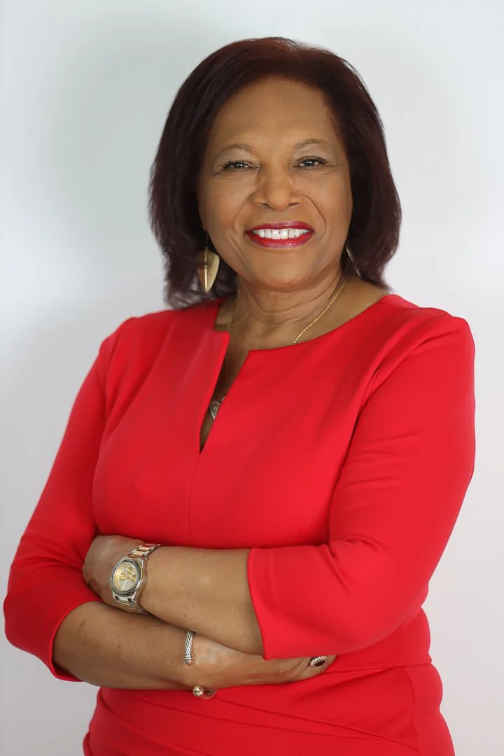 A confident woman with shoulder-length brown hair wearing a red dress, gold earrings, and a gold watch, standing with crossed arms against a white background.