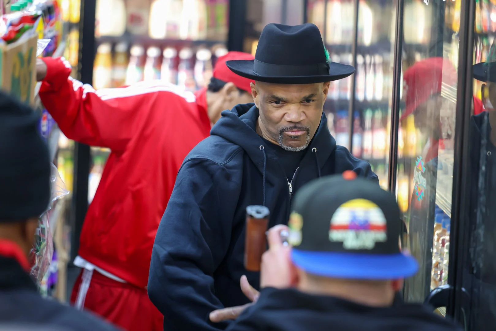 Man with a black hat and hoodie shopping at a grocery store refrigerator surrounded by people.