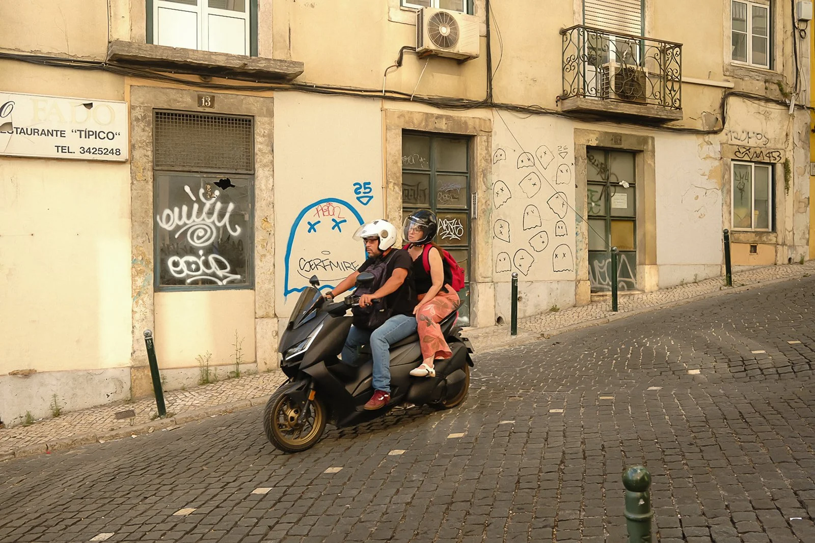 Two people riding a black scooter on a cobblestone street in front of a wall with graffiti and posters, wearing helmets and casual clothing.