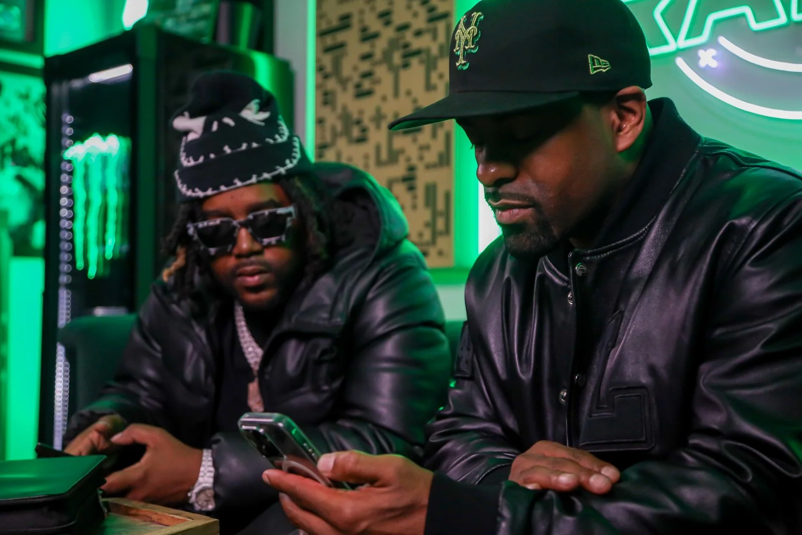 Two men in black leather jackets sitting at a neon-lit gaming or restaurant establishment, both looking at their phones. The man on the right wears a black cap with a logo, and the man on the left has dreadlocks, glasses, and a hat. Green neon lighti