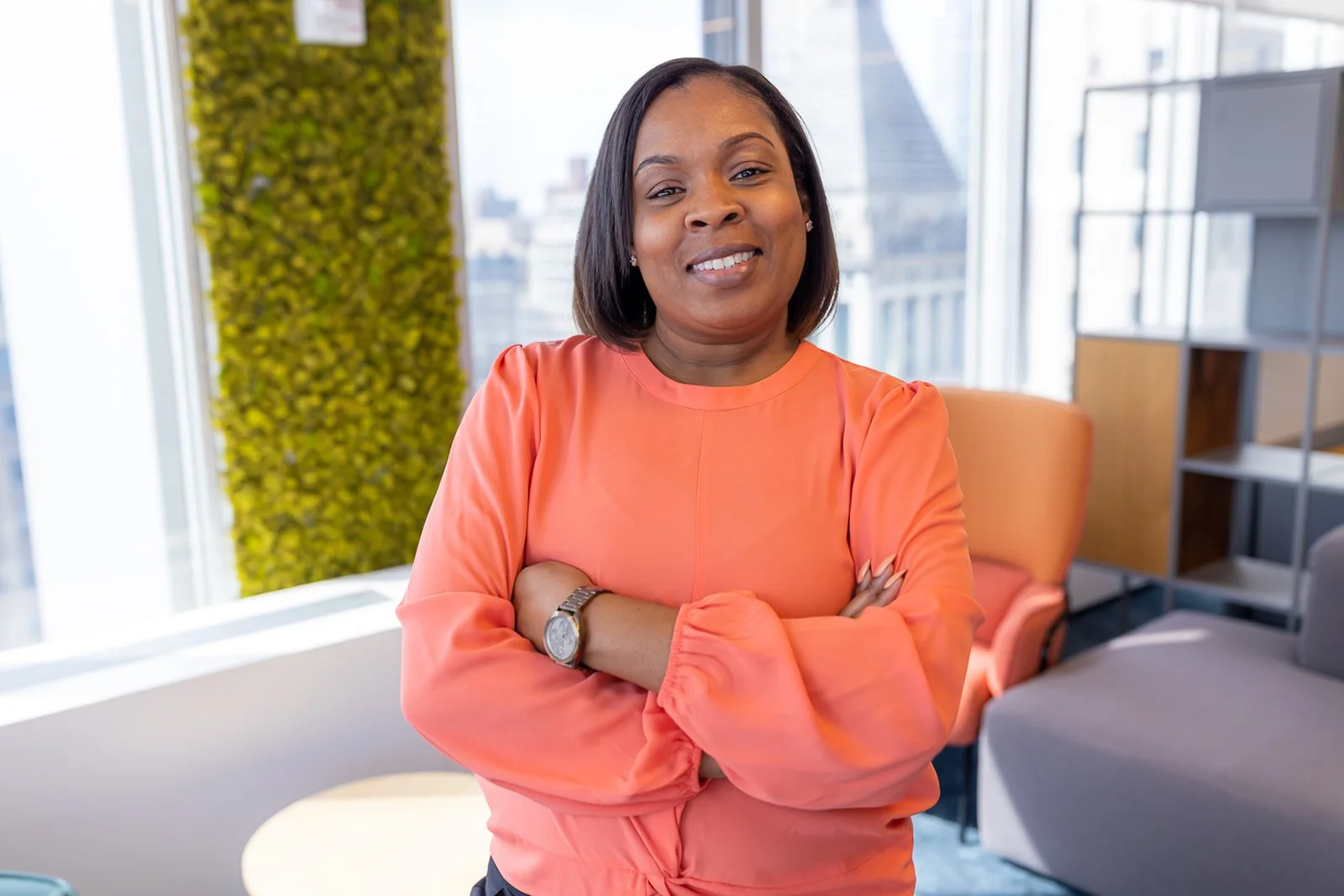 A woman in a coral long-sleeve blouse smiling with arms crossed in a modern office space, with large windows and cityscape view in the background.