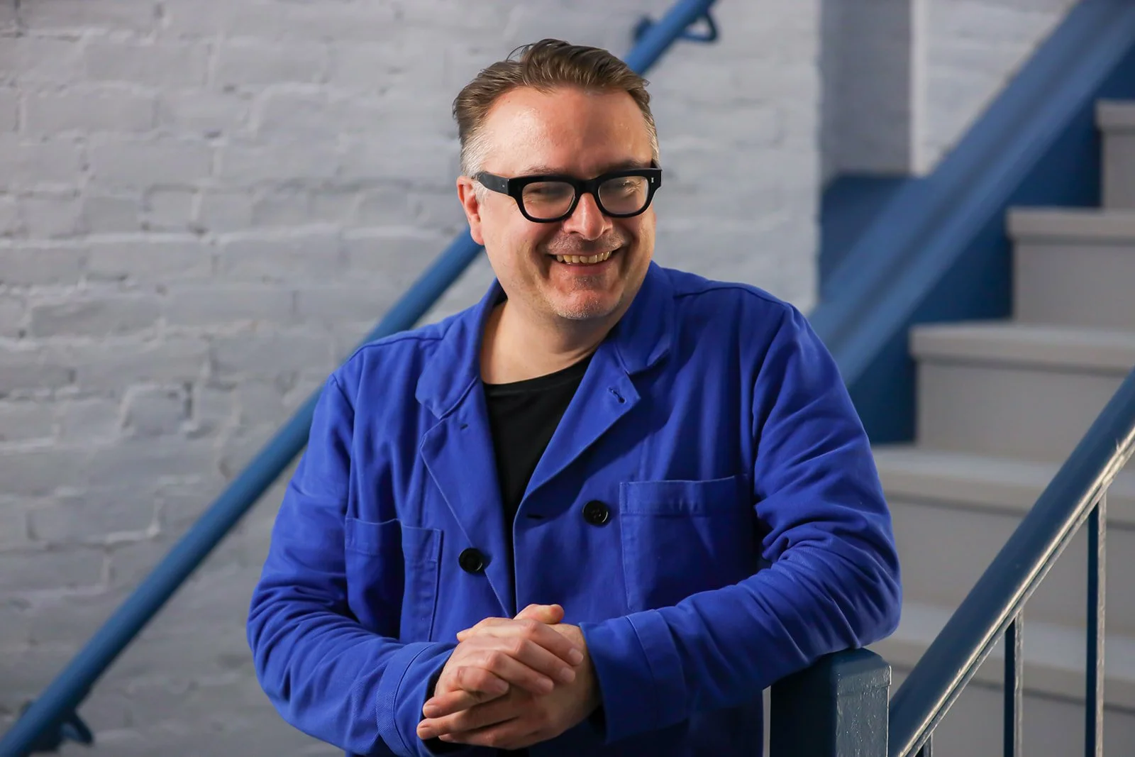A man wearing black glasses and a blue jacket standing on a staircase with gray brick walls in background.