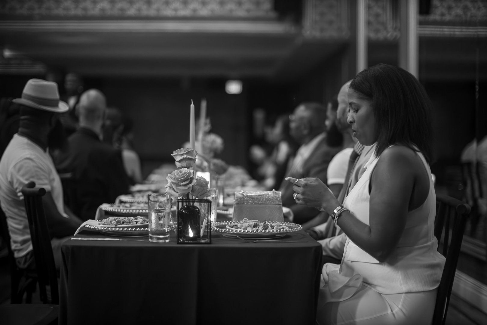 People seated at a banquet table during a formal event, with floral centerpieces, candles, and plates of food in a dimly lit room.