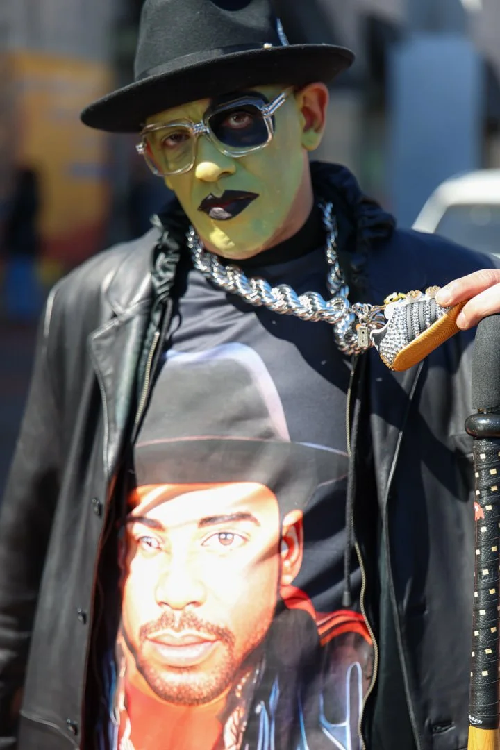 Person dressed in exaggerated makeup and accessories, wearing a black hat and leather jacket, holding a scooter handlebar, with a graphic t-shirt featuring a man's face, and a large silver chain necklace.