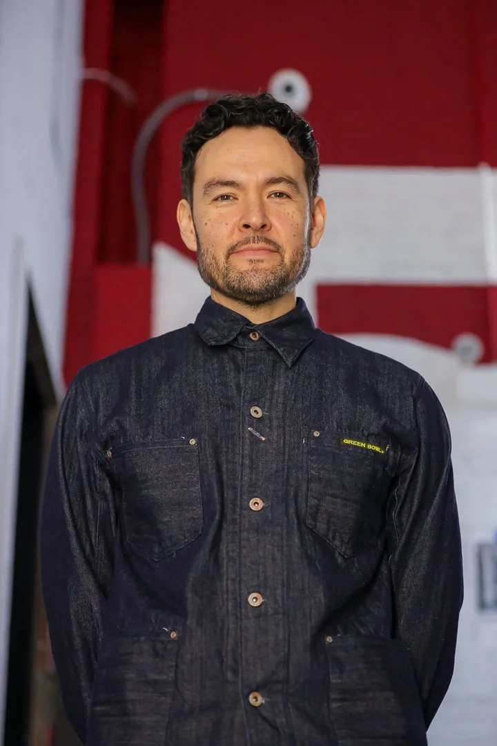 A man with dark curly hair and a beard, wearing a dark denim shirt, stands in front of a red background with white and black elements.