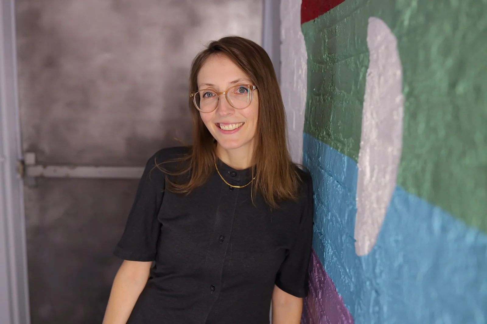 A woman with shoulder-length brown hair, wearing glasses and a black top, smiling and leaning against a colorful wall with a smiling face painted on it.