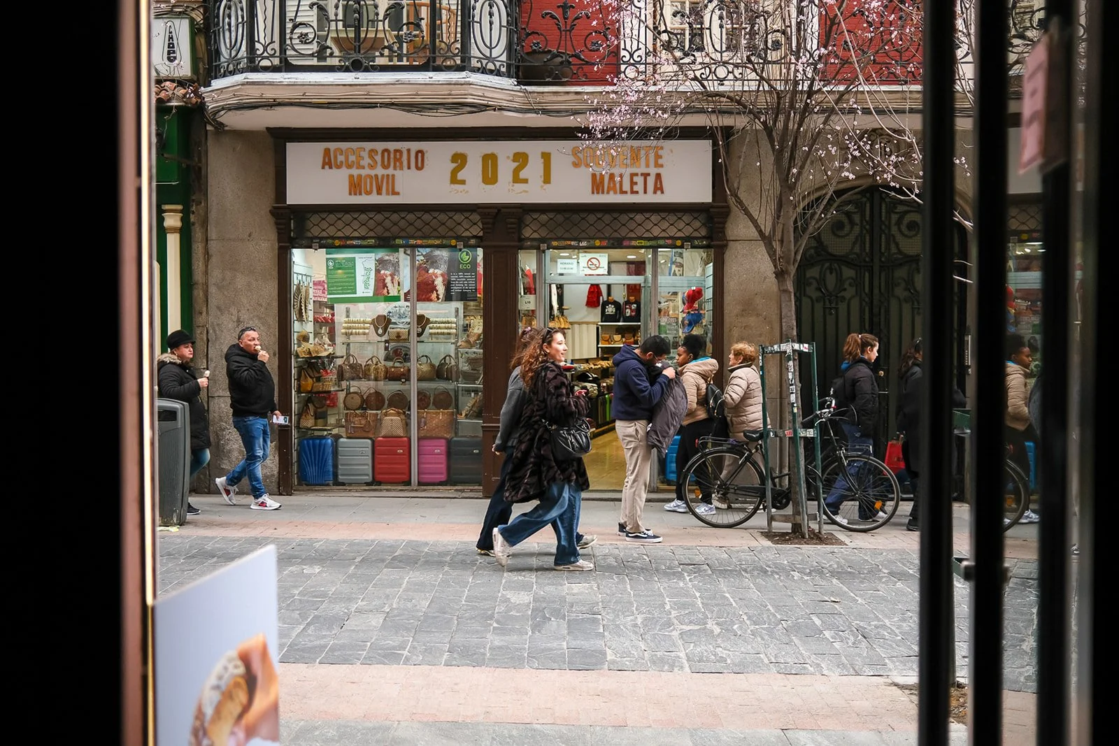 Street scene viewed through a window with black bars, showing people walking past a store with a sign that reads "Accesorio Movil 2021 Solvente Maleta." Several individuals are walking and standing outside, and bicycles are parked nearby, with a tree