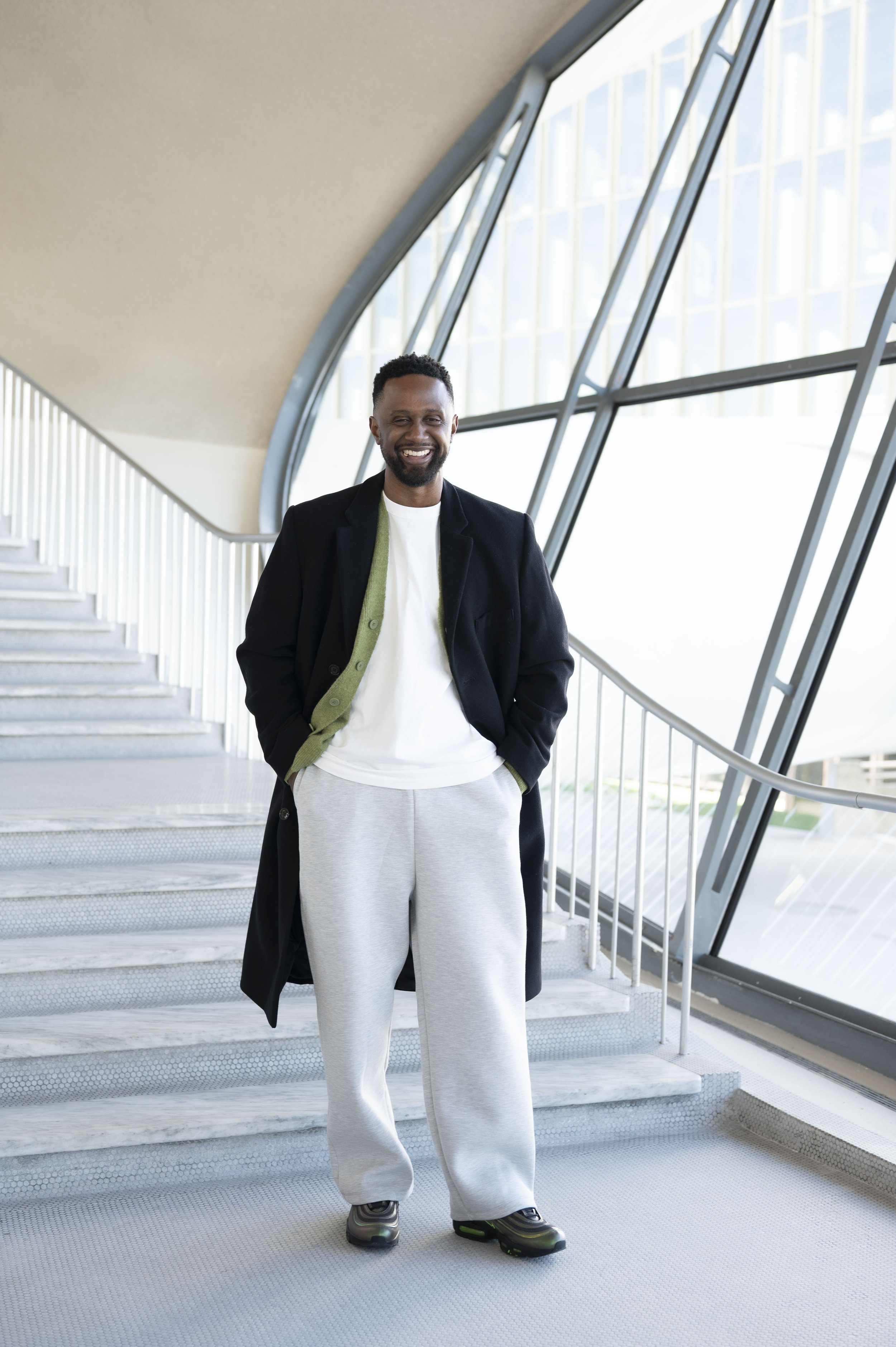 A smiling man standing on stairs in a modern building near large glass windows, wearing a black coat, white shirt, gray sweatpants, and sneakers.