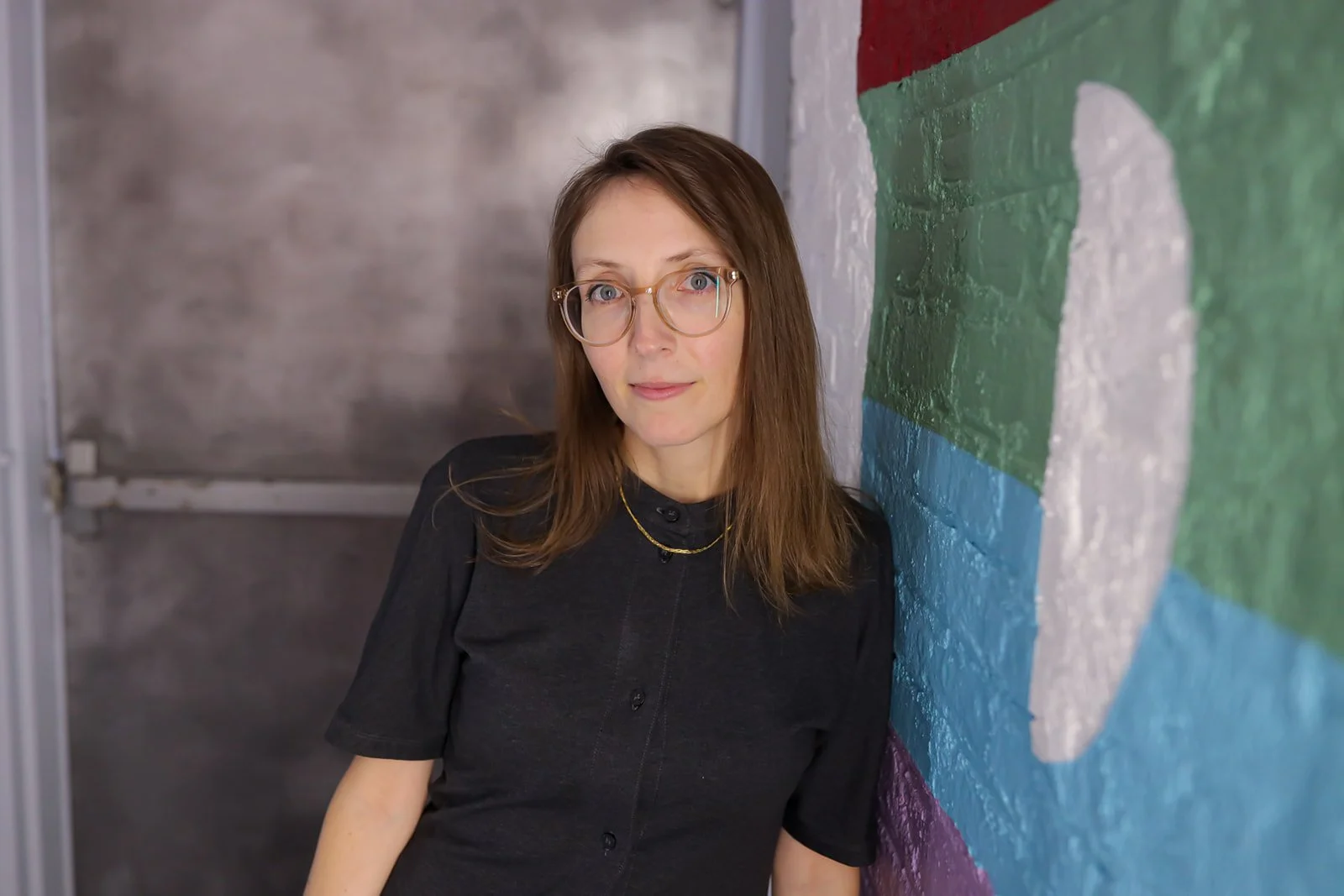 A woman with long brown hair and glasses, wearing a black shirt with a gold necklace, leaning against a painted brick wall.