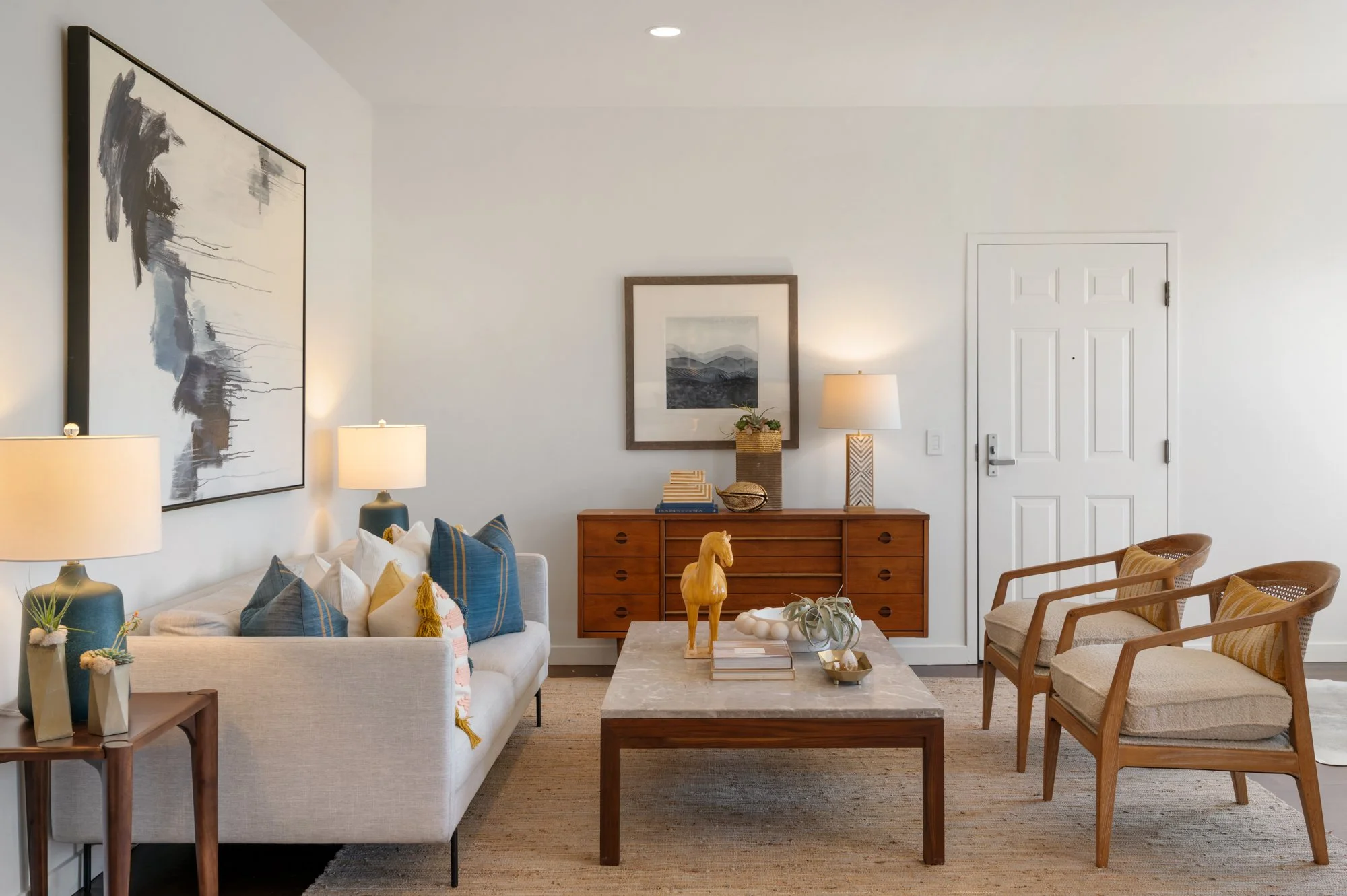 Living room with a white sofa decorated with blue and yellow pillows, a marble coffee table with decorative objects, and wooden chairs with beige cushions. The wall has framed artwork, and there are two table lamps with white shades. There is a woode