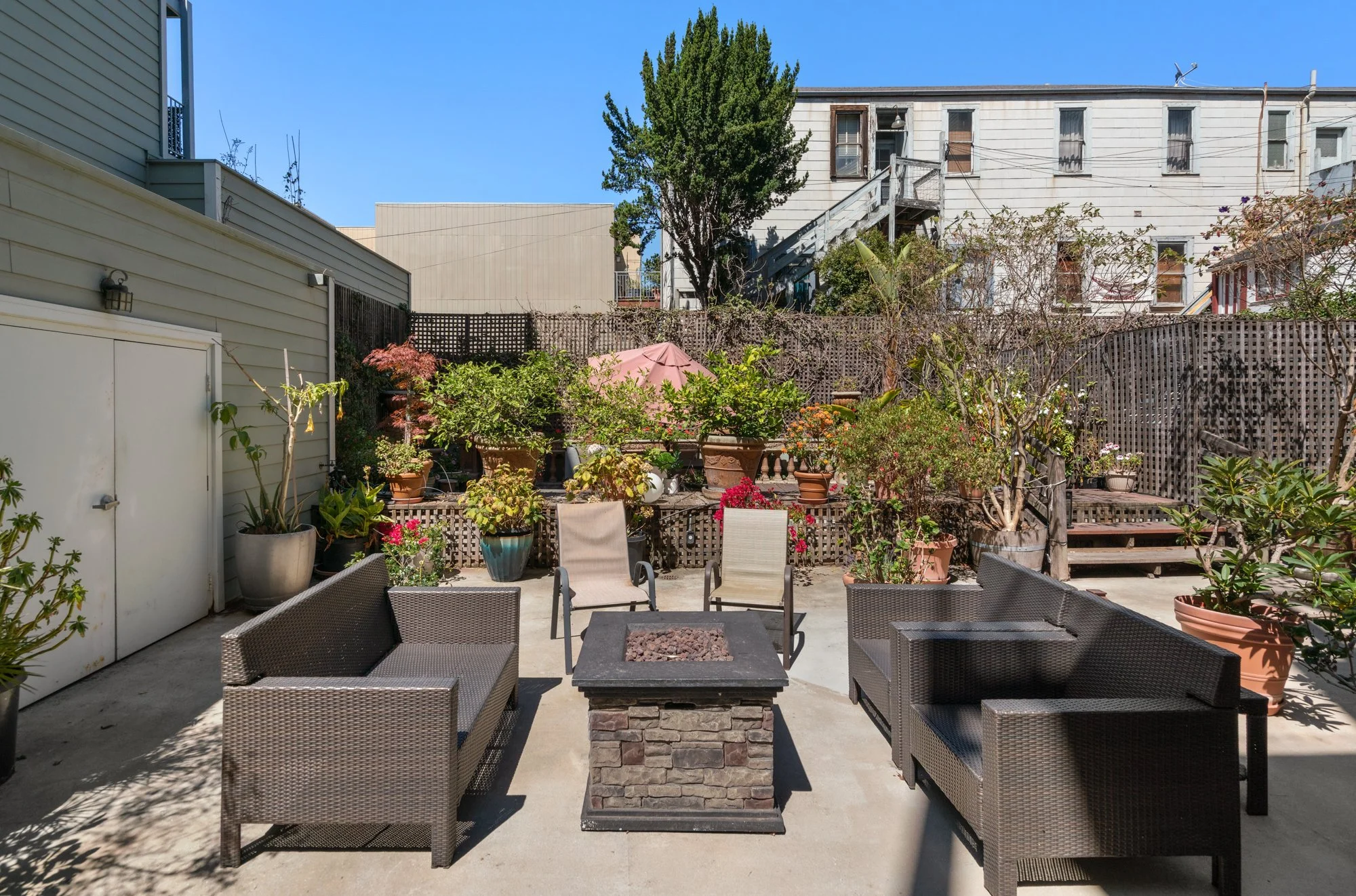 A backyard patio with outdoor seating, potted plants, and a central fire pit, enclosed by a wooden fence and neighboring houses with stairs and trees visible in the background.