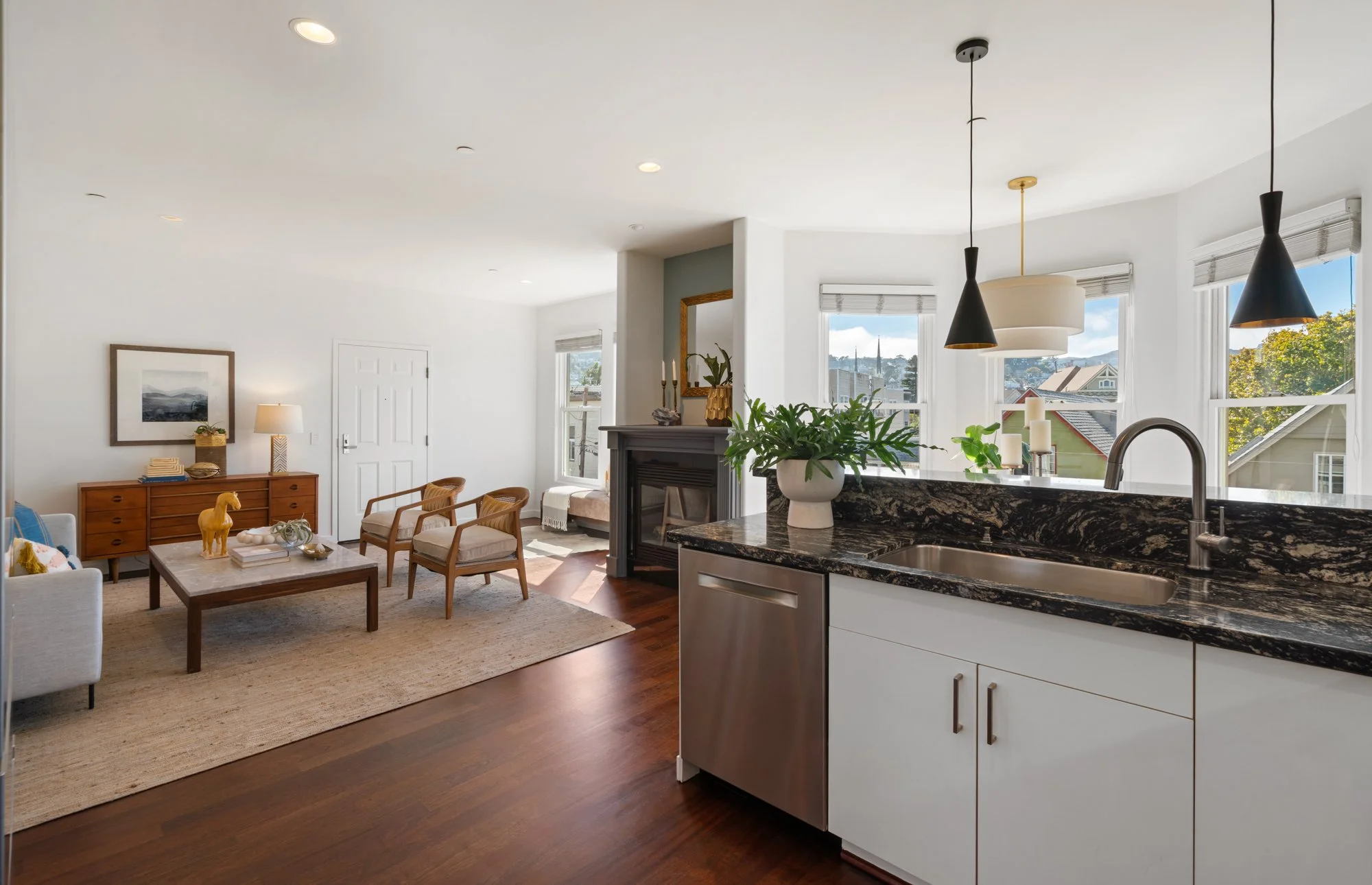 Modern living room and kitchen with white walls, hardwood floors, and large windows allowing natural light, featuring framed artwork, a fireplace, green plants, wood furniture, and black pendant lights.