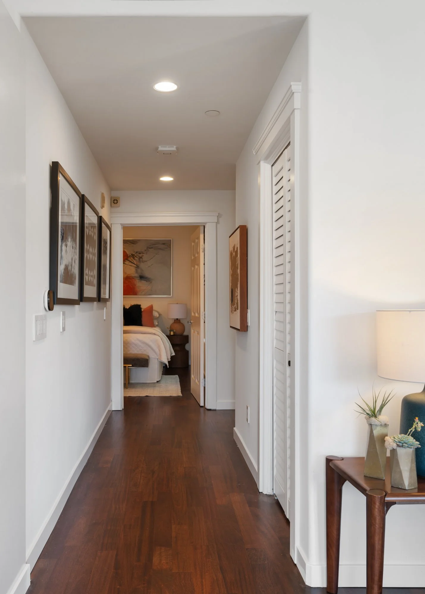 Hallway leading to a bedroom, with dark hardwood flooring, white walls, framed artwork on the left, a side table with decor on the right, and a partially visible folded louvered closet door.