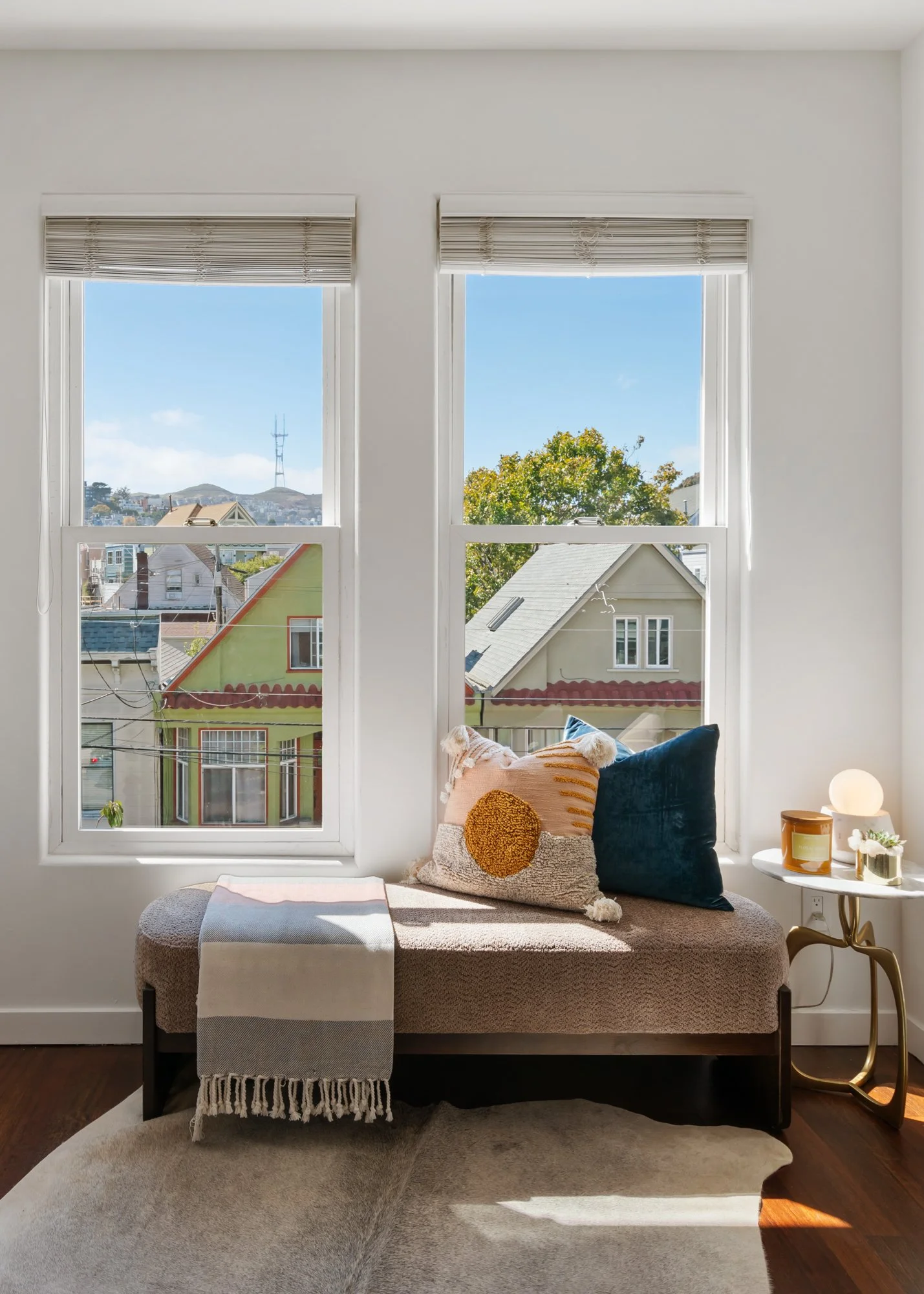 A cozy window seat with throw pillows, a beige blanket with fringe, and a small table with decorative items, overlooking rooftops and trees outside during daytime.