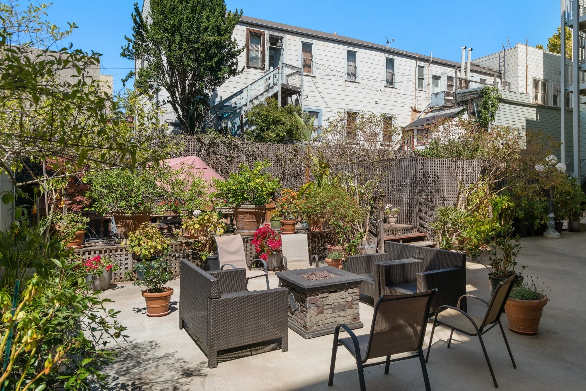 Backyard patio with outdoor furniture, potted plants, and a small fire pit, enclosed by a wooden fence, with a two-story white house with weathered siding and large windows in the background under a clear blue sky.