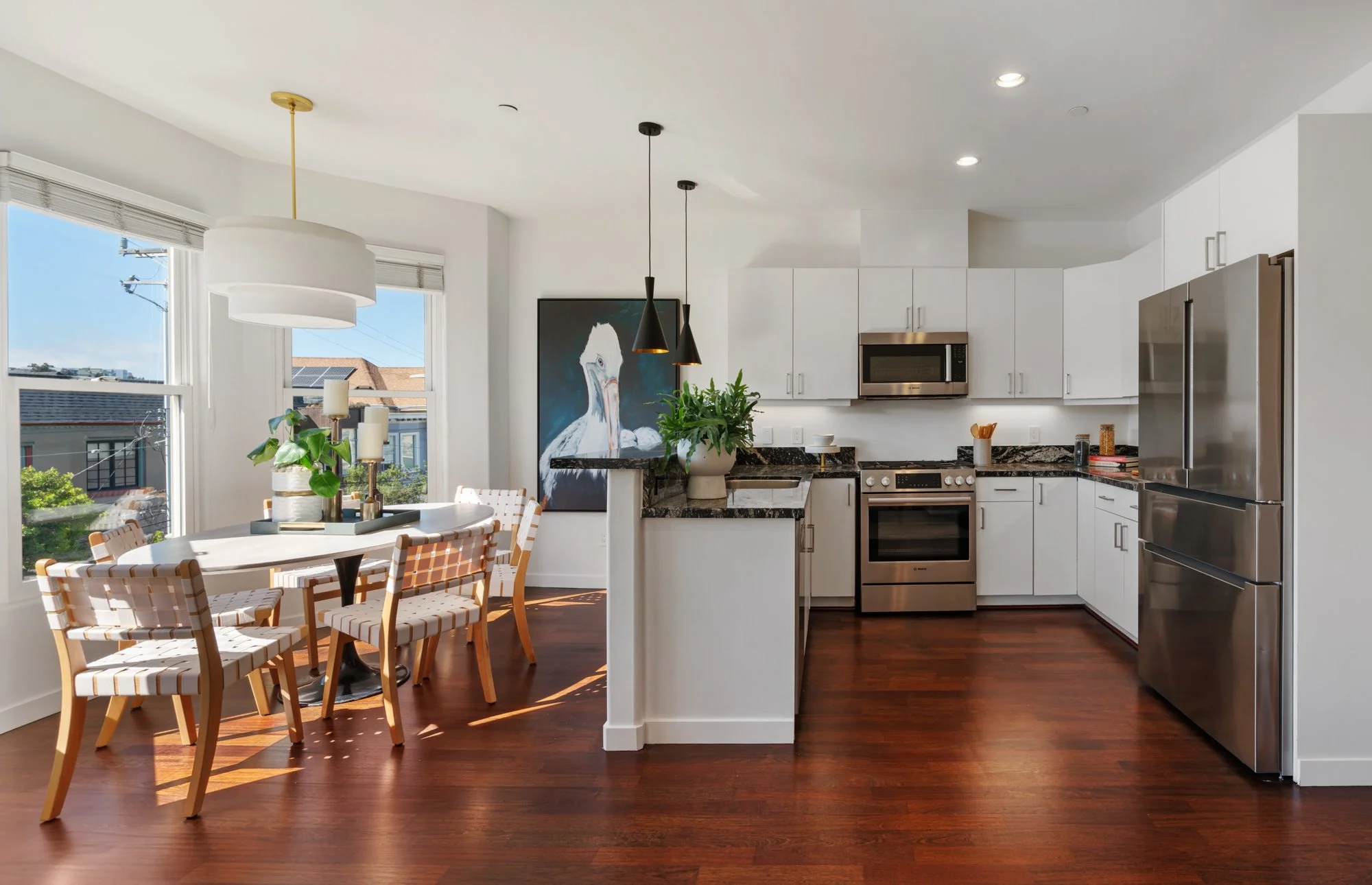 Bright open-concept kitchen and dining area with white cabinetry, stainless steel appliances, a black and white marble countertop, and a dining table with patterned chairs. Large windows let in natural light, and a large framed picture of a swan deco