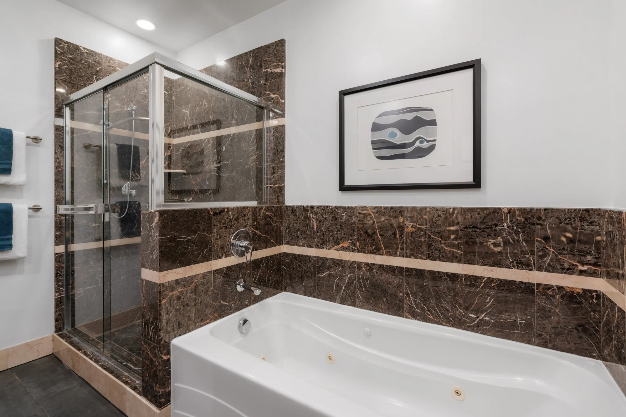 Bathroom with brown marble walls and glass shower enclosure, a white bathtub with jets, a framed abstract art piece, and towels on a towel rack.
