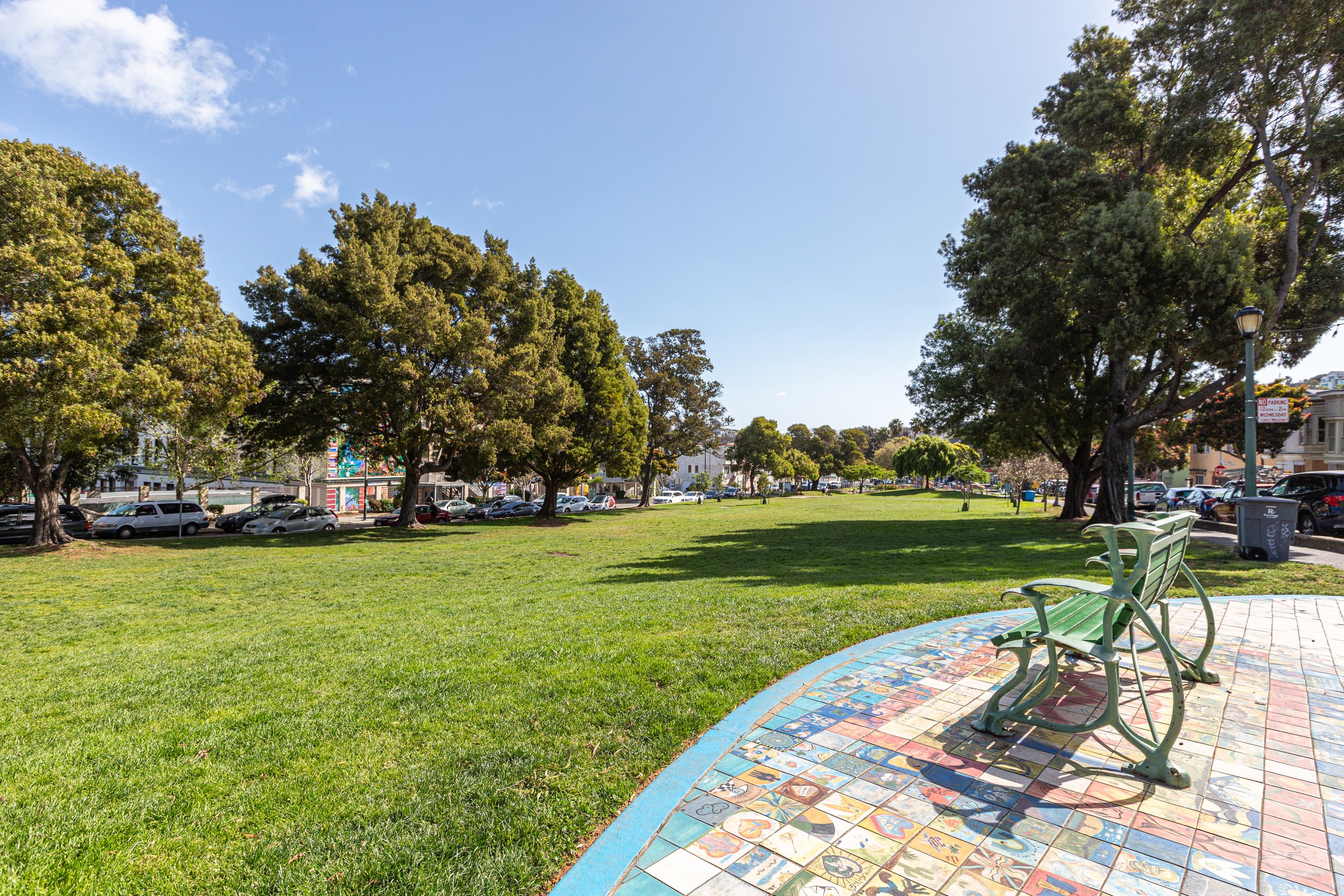 A park with green grass and tall trees under a clear blue sky. There are parking spaces filled with cars along the street, benches, and a colorful mosaic sidewalk.