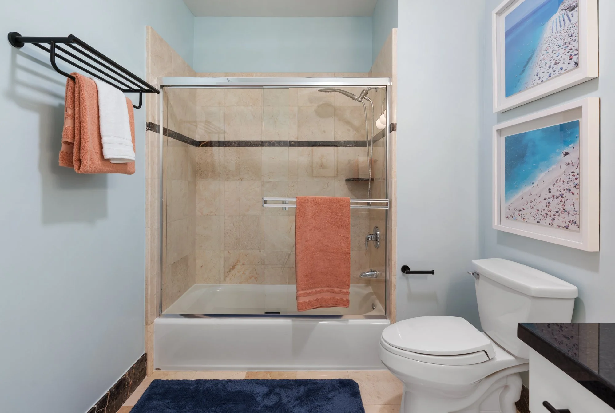 Bathroom with a glass-enclosed shower, toilet, white walls, three beach-themed art prints, a black towel rack with pink and white towels, and a navy blue rug.