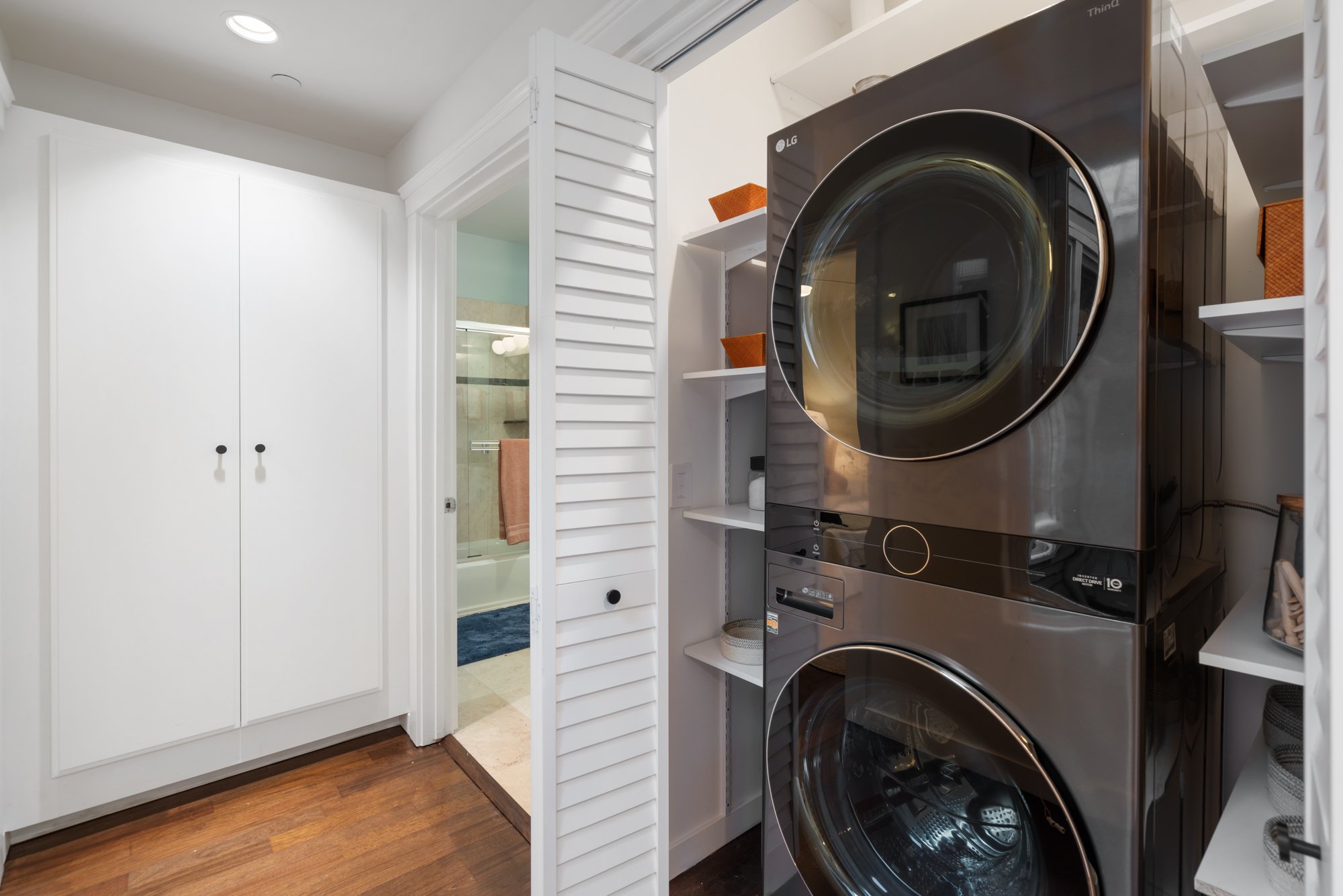 Front view of a laundry room with a black washer and dryer, white shelving, two orange storage baskets, a white closet with two doors, and a doorway to a bathroom with a glass shower door and orange towel.