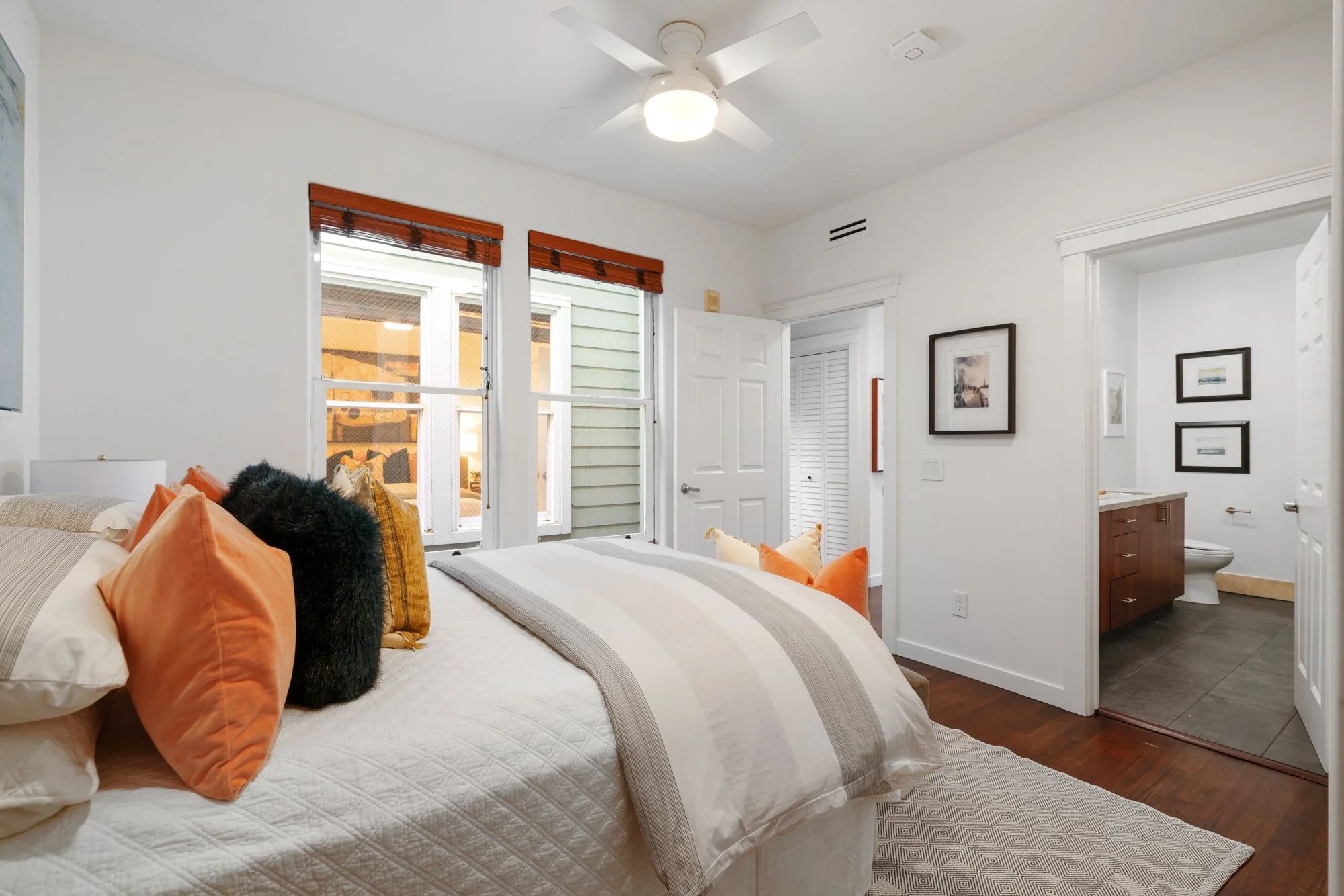 A bedroom with a bed covered in white bedding with beige and orange pillows. Two windows with wooden blinds are on the wall, allowing natural light. A ceiling fan is mounted on the ceiling, and a door leads to a bathroom with a vanity and a toilet. T