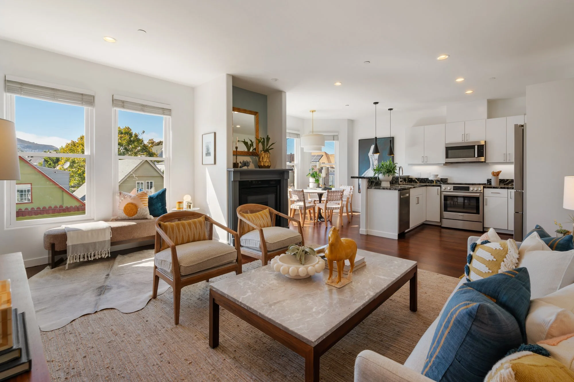 Bright living room with large windows, beige sofa, wooden chairs, fire place, round coffee table with decorative objects, and an open kitchen with white cabinets and black countertops.