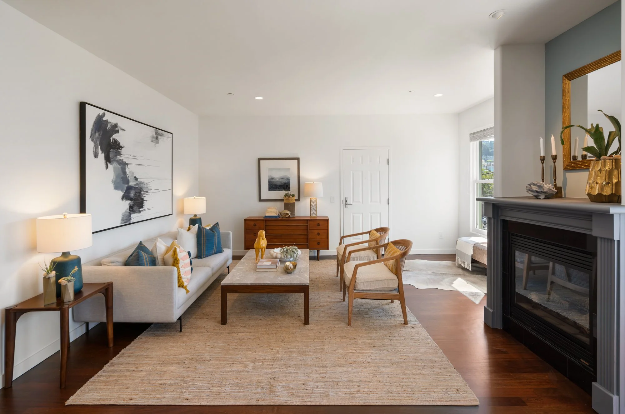 Living room with white walls, wooden furniture, modern art, a fireplace, and large windows letting in natural light.