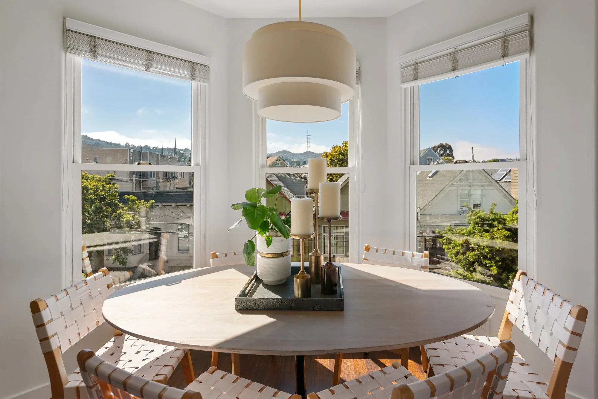 Dining area with a round table and woven chairs near large windows with a cityscape view and blue sky.