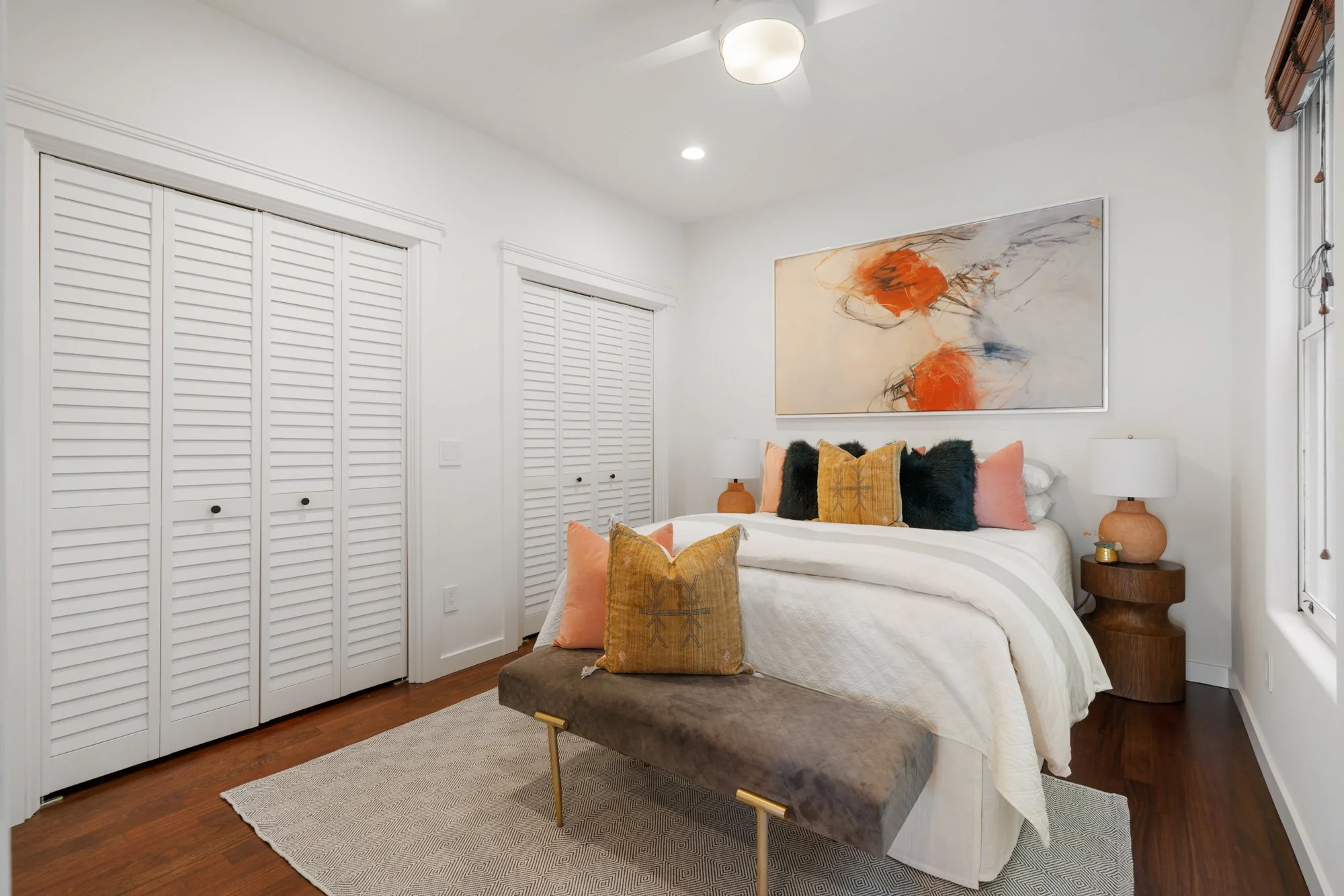 A cozy bedroom with a white bed, colorful pillows, and a wooden bedside table with a lamp. Abstract art hangs above the bed, and there are white closet doors and a window with wooden blinds in the room.