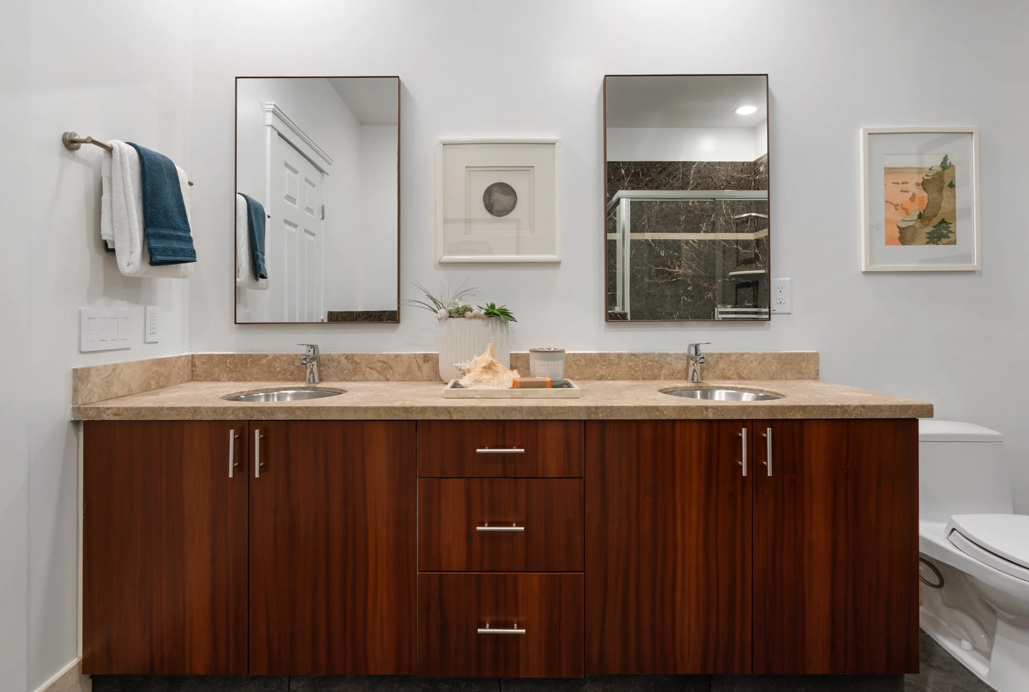 A modern bathroom vanity with a marble countertop, two sinks, and twin mirrors above, with towels hanging on the wall and framed artwork on the wall.