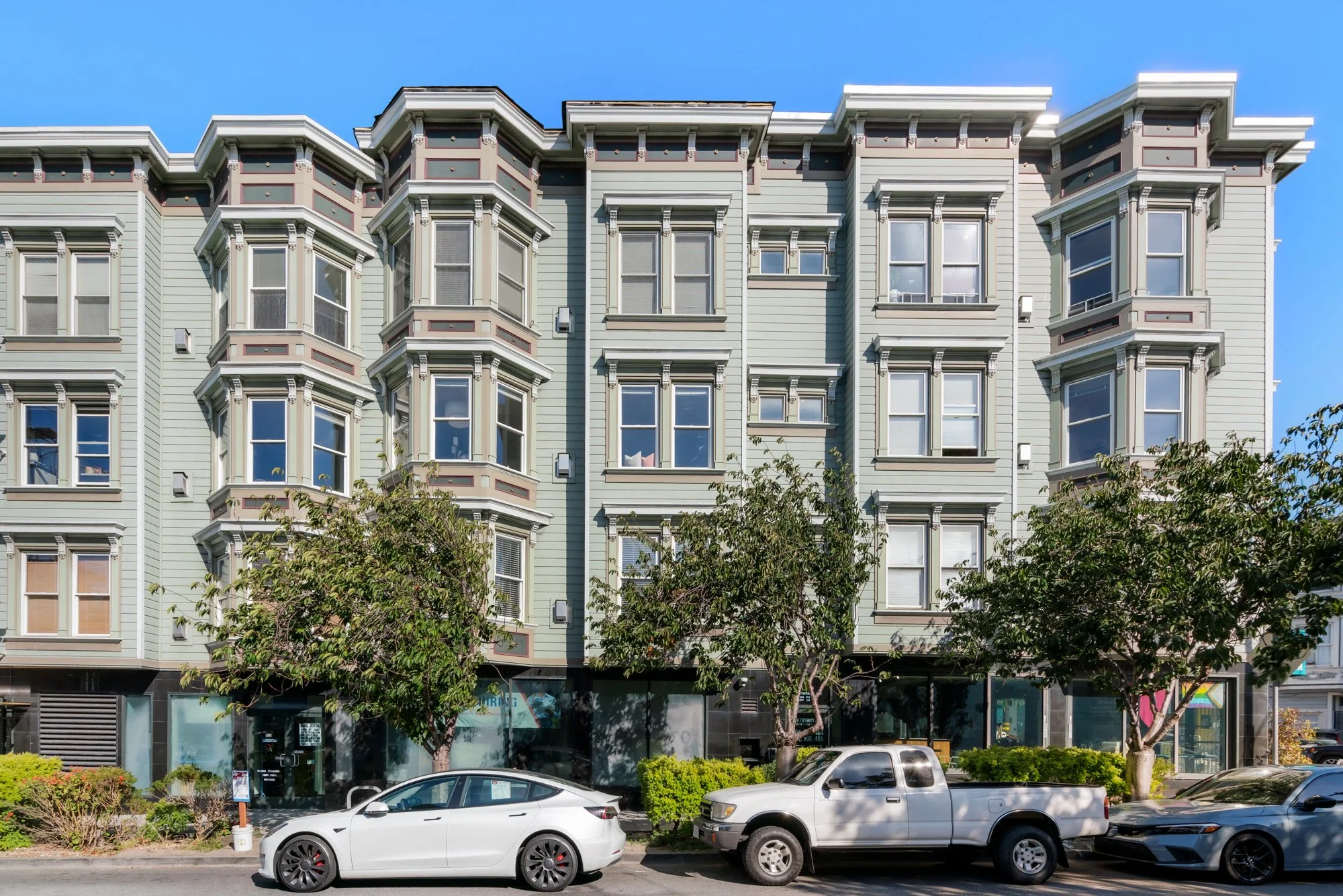 A multi-story apartment building with light green siding, bay windows, and ornate architectural details, situated on a city street with parked cars and trees in the foreground.