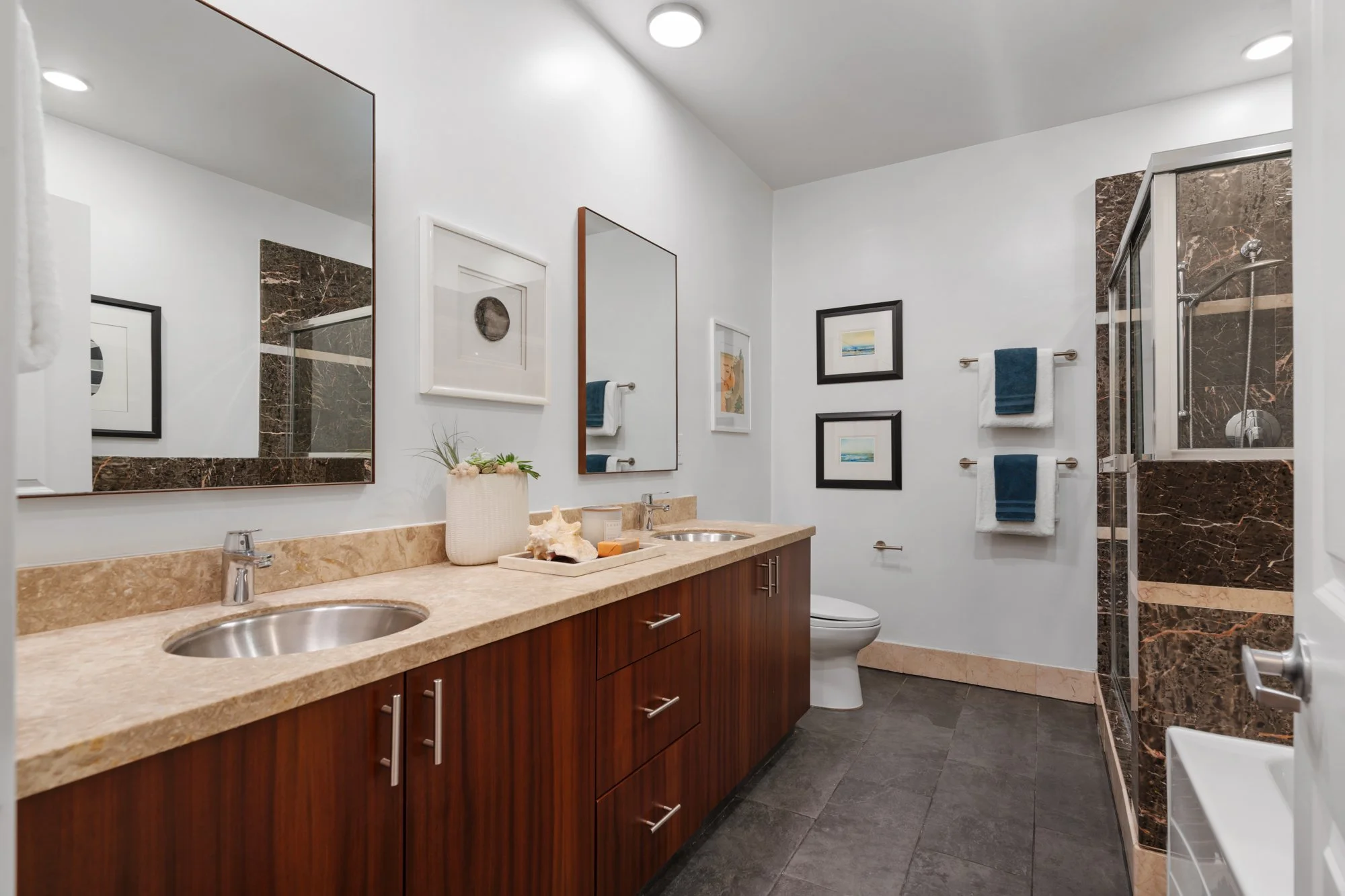 Bathroom with dual sinks, large mirrors, framed artwork on white walls, brown marble accents, towels hanging on a silver rack, and a glass-enclosed shower.