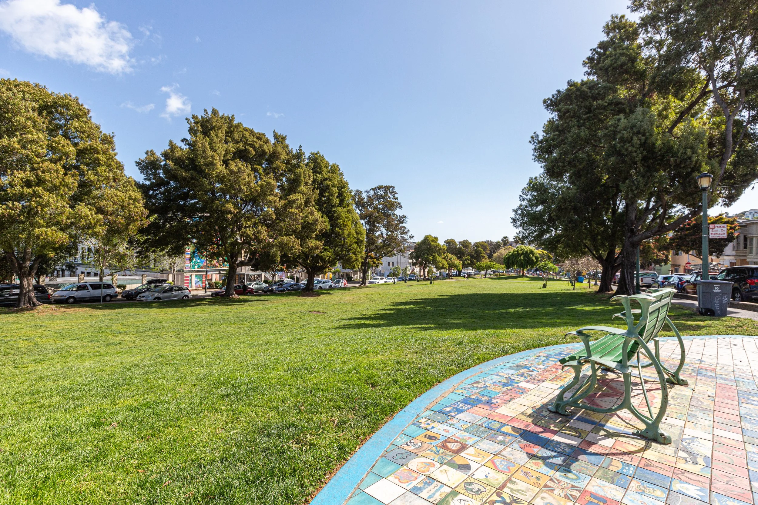 A park with green grass, large trees, and a colorful tiled pathway with green benches, park signage, and parked cars in the background under a clear blue sky.