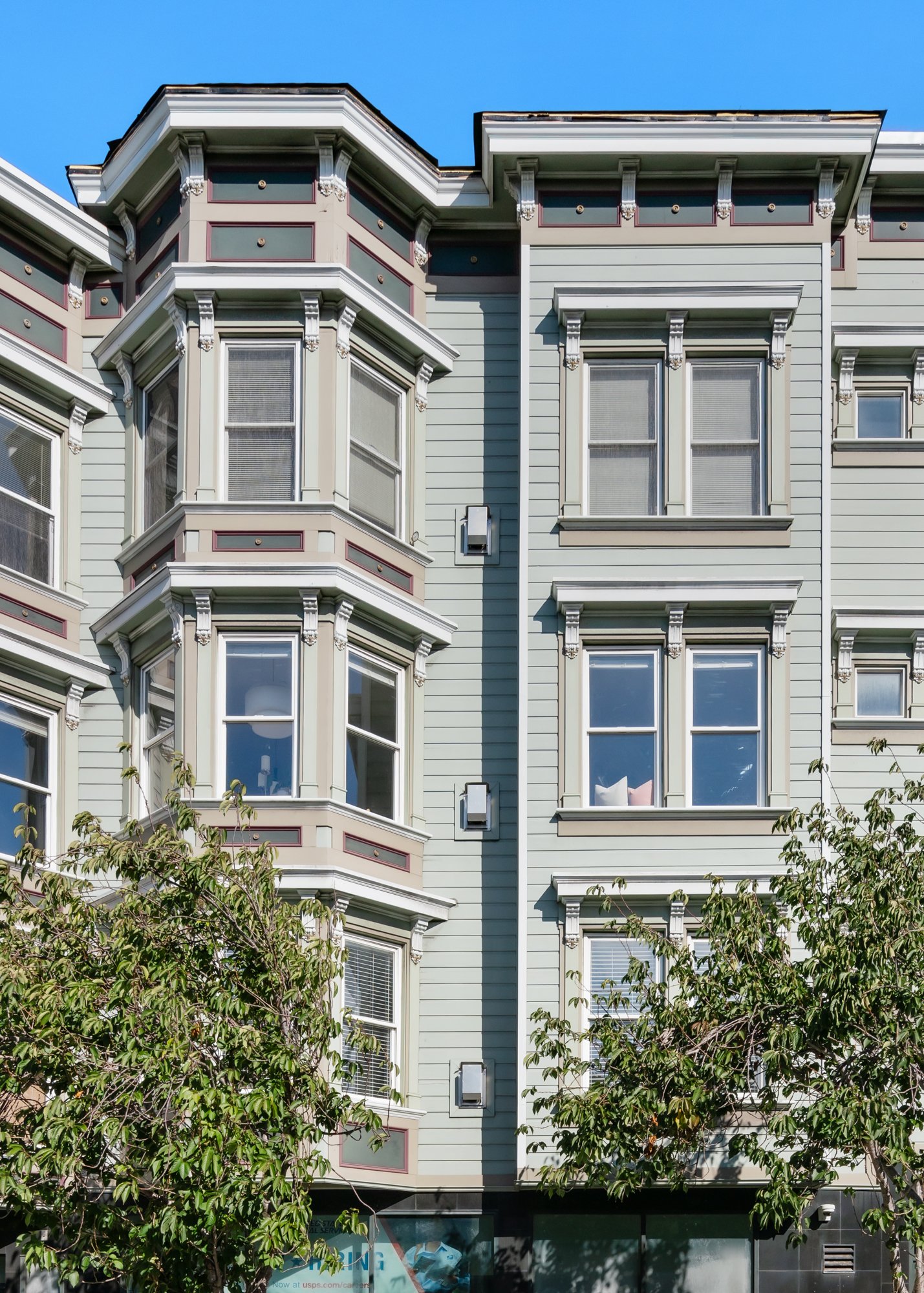 Exterior view of a Victorian-style apartment building with bay windows, light pastel green siding, white trim, and decorative cornices, with trees in the foreground and a bright blue sky.