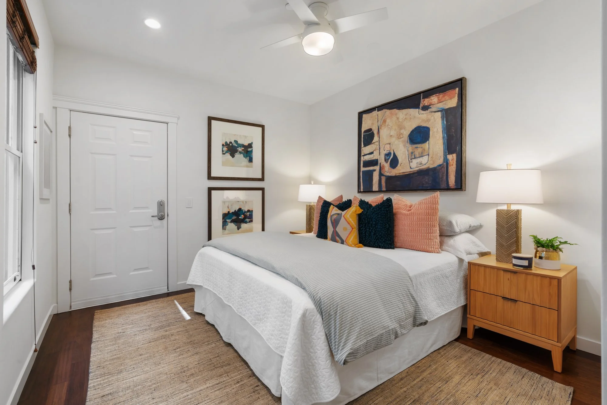 A bedroom with a white bed, colorful pillows, wooden nightstand, white lamps, and framed artwork on white walls.