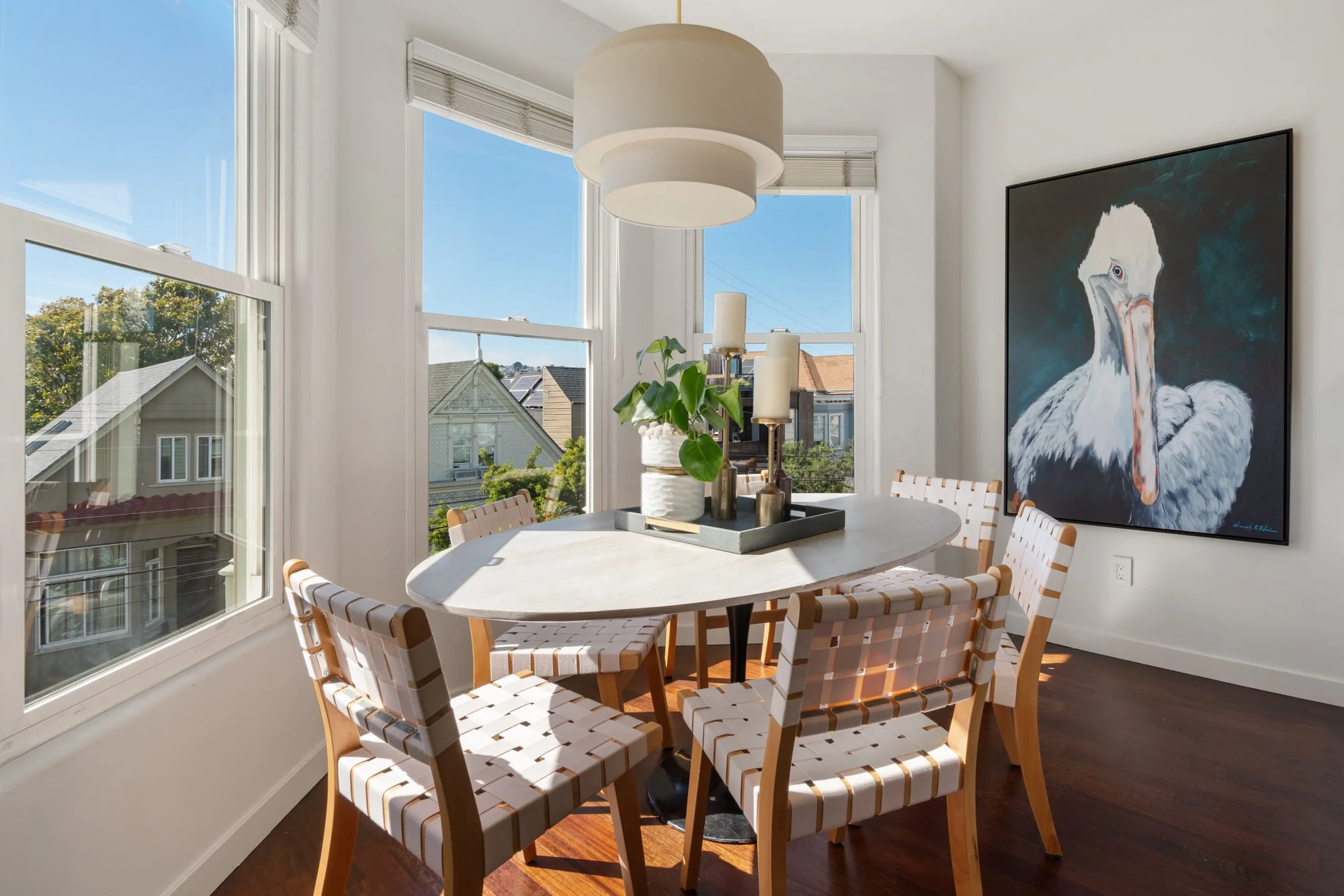 Bright dining room with a round table, six striped chairs, large windows with a view of neighboring houses and trees, modern pendant light, and a large abstract bird painting on the wall.