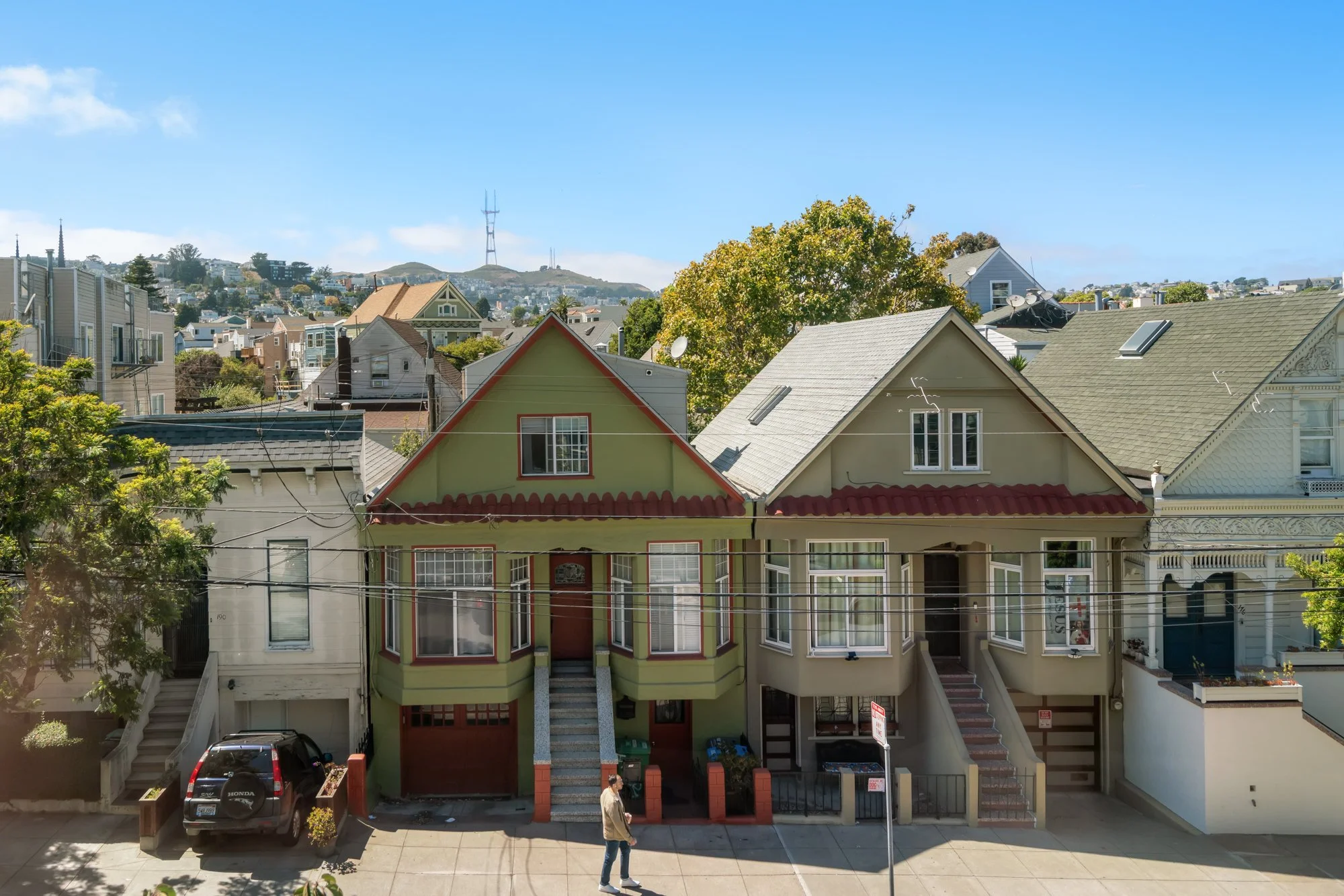 Residential street with colorful houses, some with elaborate trim, and cars parked in front. People walking on the sidewalk, trees in the background, and hills with a radio tower visible against a bright blue sky.
