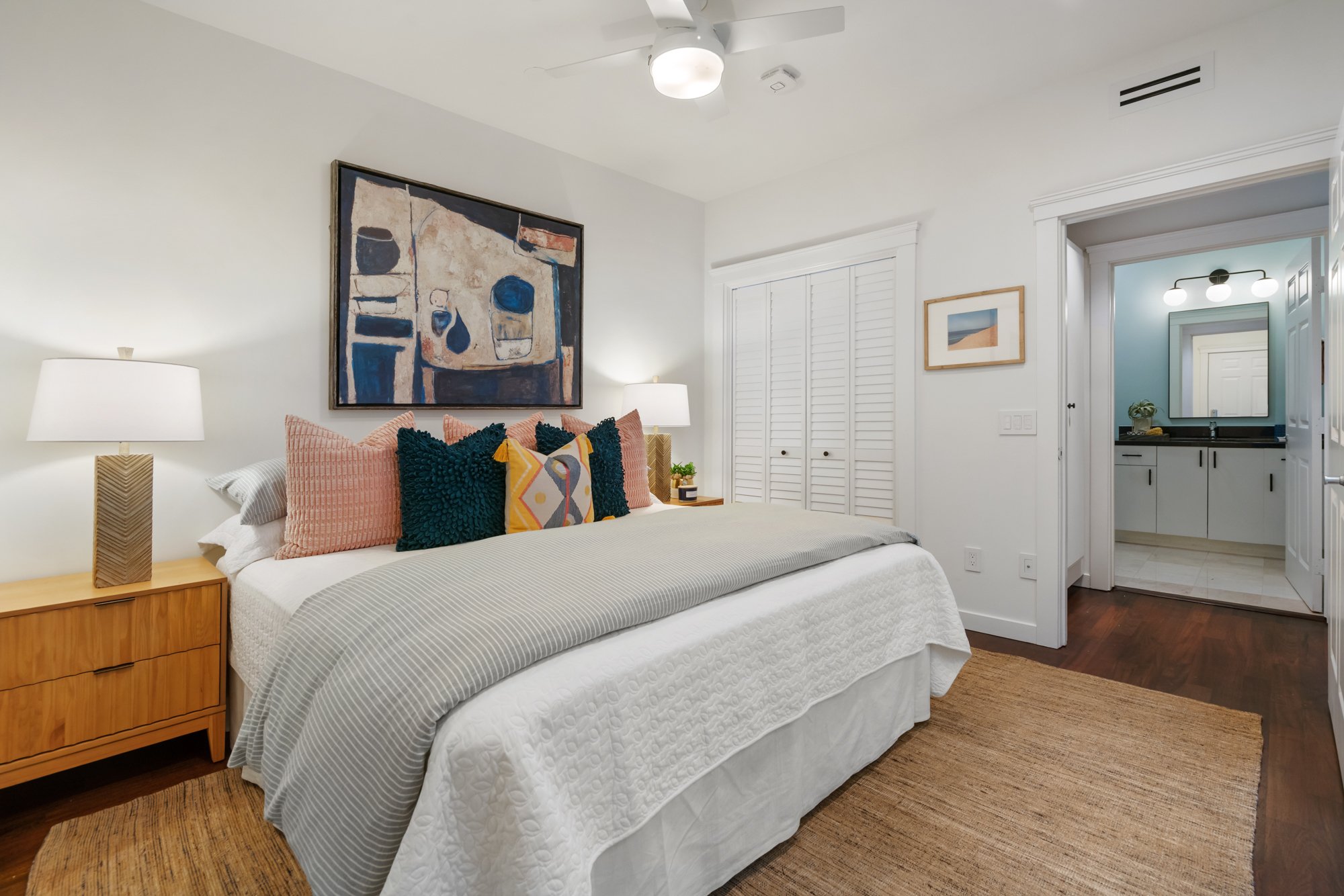 A bedroom with a white bedspread, multiple decorative pillows including dark blue, pink, and patterned ones, a wooden bedside table with a lamp, a framed abstract painting above the bed, a small bedside plant, and a closet with louvered doors. Adjace