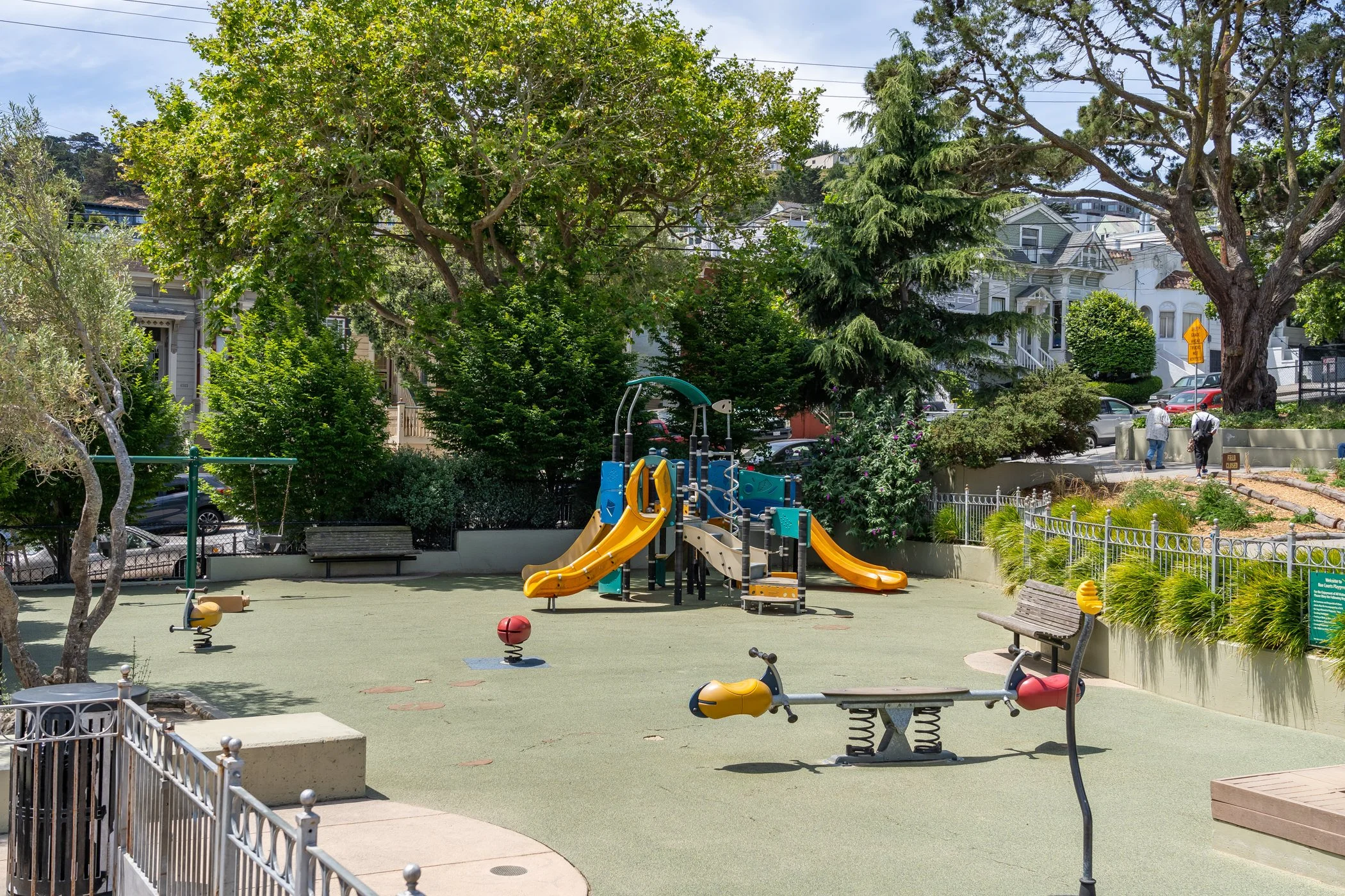 A playground with slides, spring riders, and other equipment, surrounded by green trees and residential houses in the background.