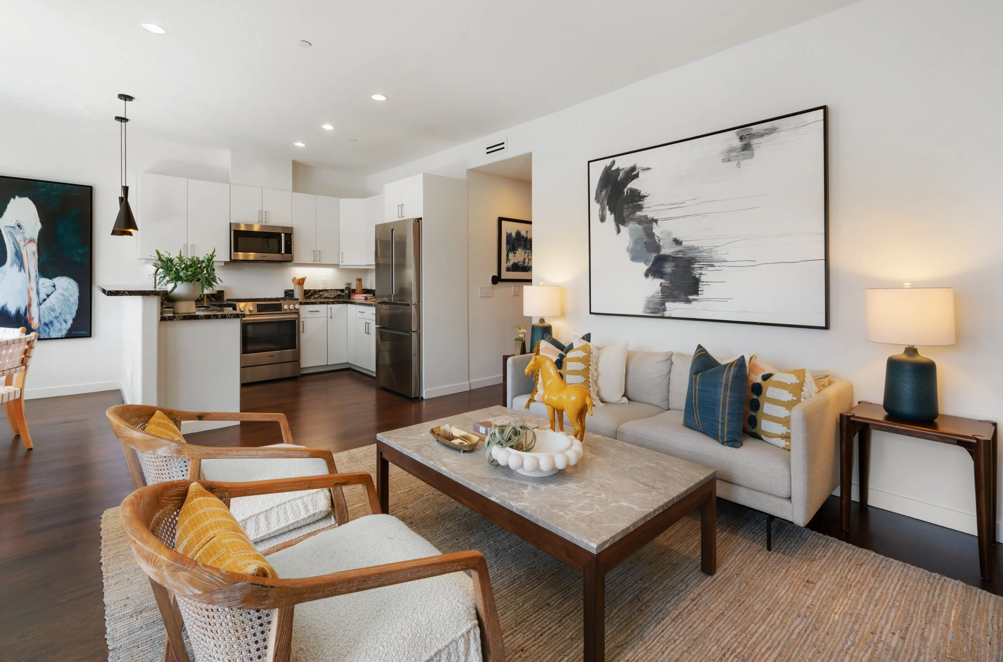 Modern living room with a gray sofa accented with patterned pillows, a marble coffee table with decorative items, and a large abstract black and white artwork on the wall. Adjacent open kitchen features white cabinets, stainless steel appliances, and