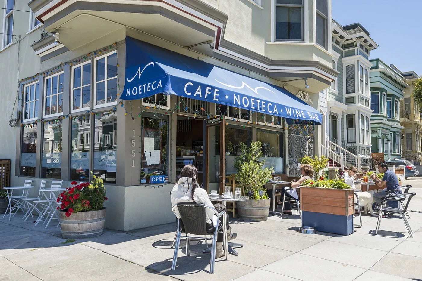 Exterior of a café named Noeteca with outdoor seating, patrons dining, plants, and a blue awning that says 'CAFE NOETEA WINE BAR' on a sunny day.